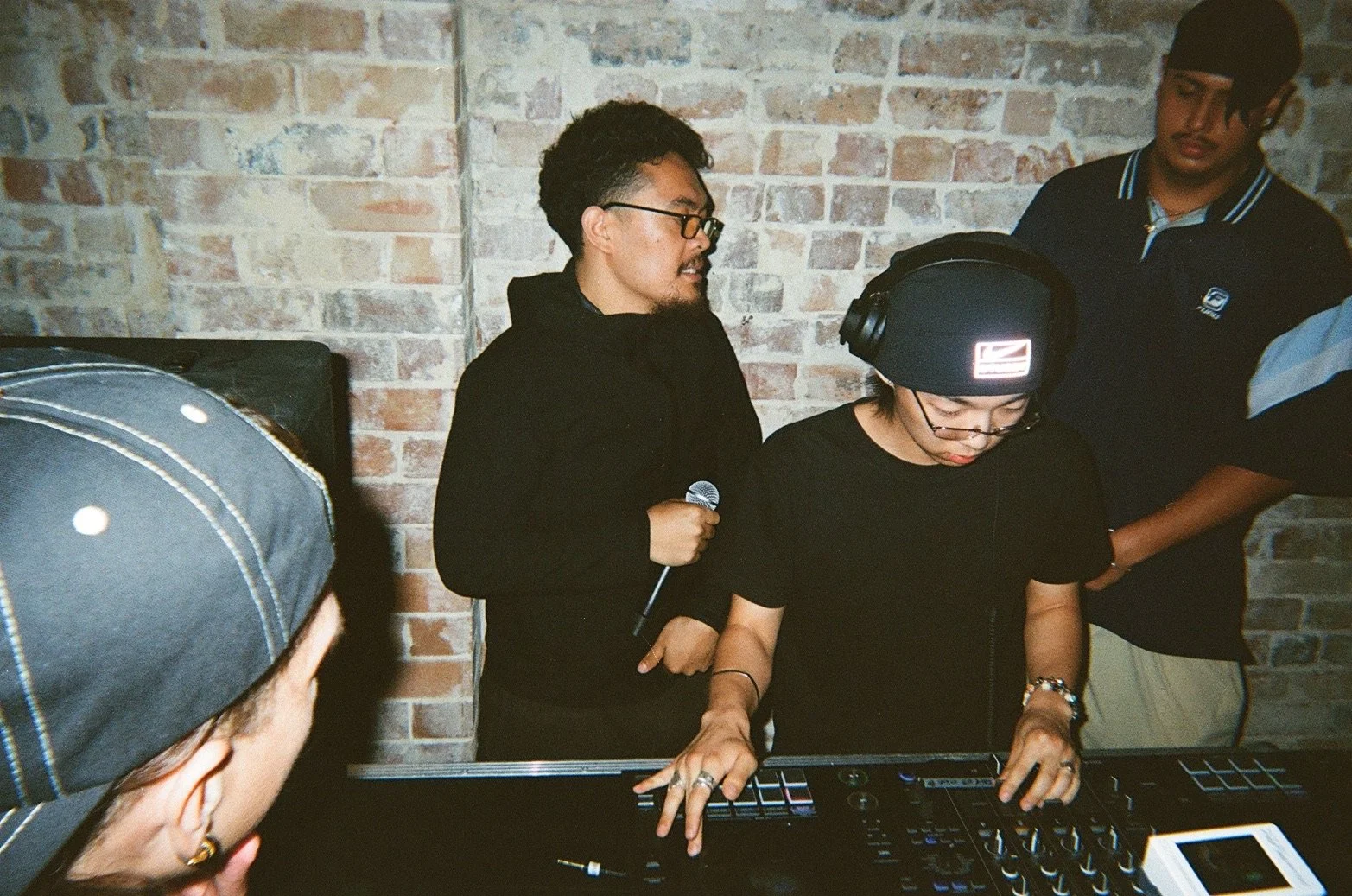 A woman wearing a black beanie, glasses, and black shirt is using a music production pad at a DJ setup. A man with glasses and a black hoodie is standing behind her, holding a microphone, and another person in a black jacket and a person in a police-