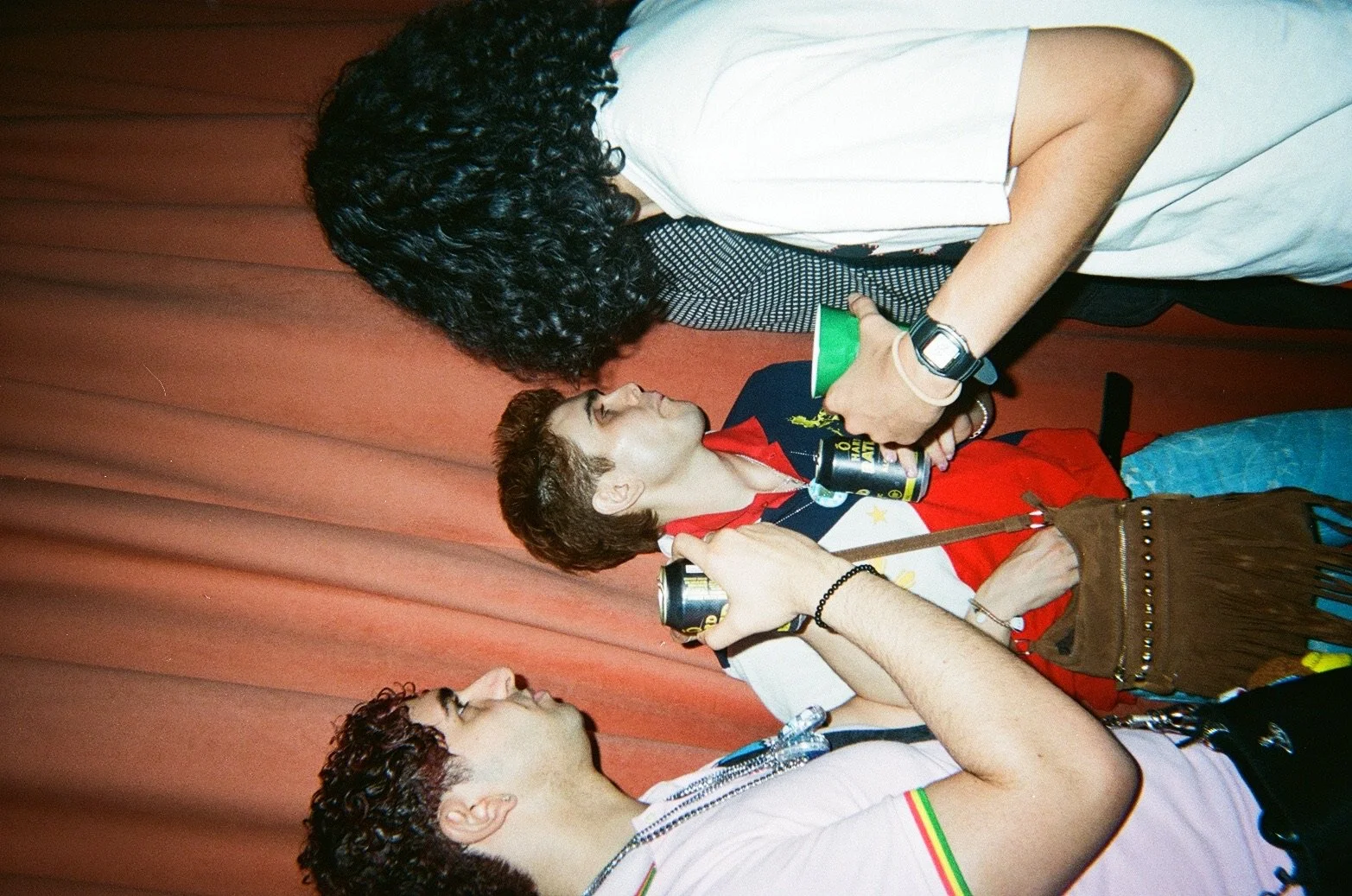 Three young people at a social gathering, one standing and two sitting, holding cans, with conversations taking place.