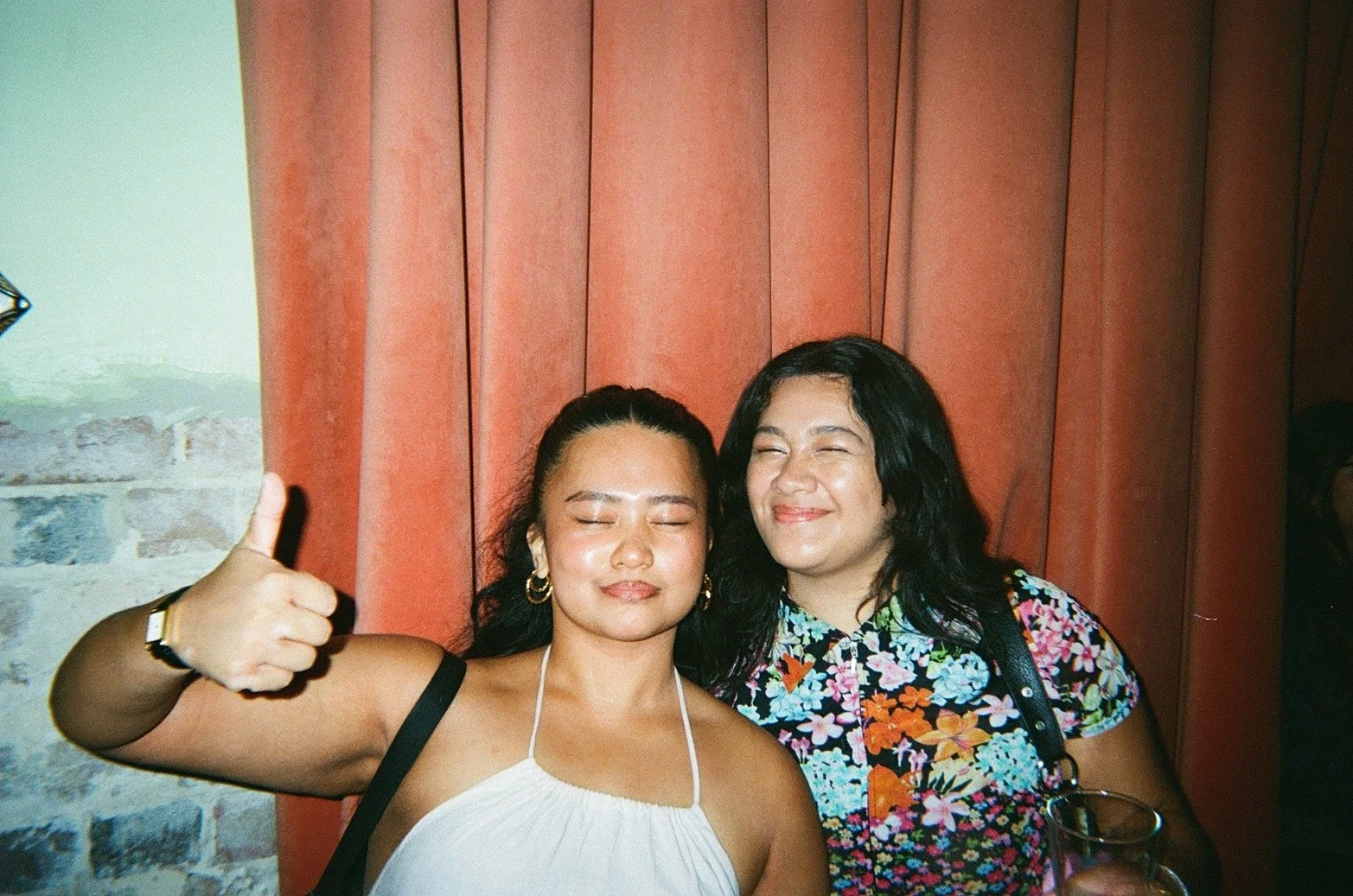 Two women with closed eyes smiling and posing for a photo with one giving a thumbs up. They are standing in front of a red curtain, one wearing a white dress and the other wearing a colorful floral shirt, holding a glass of beverage.