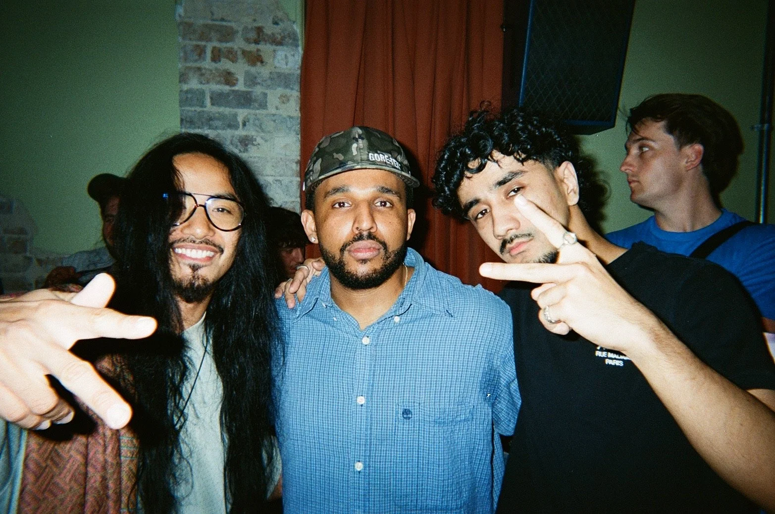 Three men posing together indoors, with one making a peace sign and another pointing toward the camera, in a casual social setting.