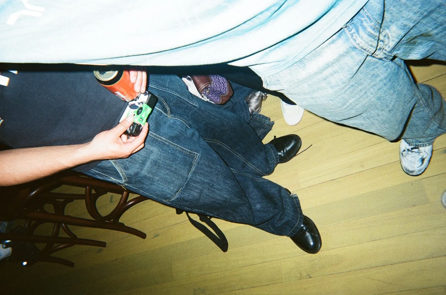 Overhead view of people standing in line, showing their legs, feet, and part of their torsos. One person is holding a green lighter and a soda can, another has a purse, and they are wearing various shoes including boots and sneakers.