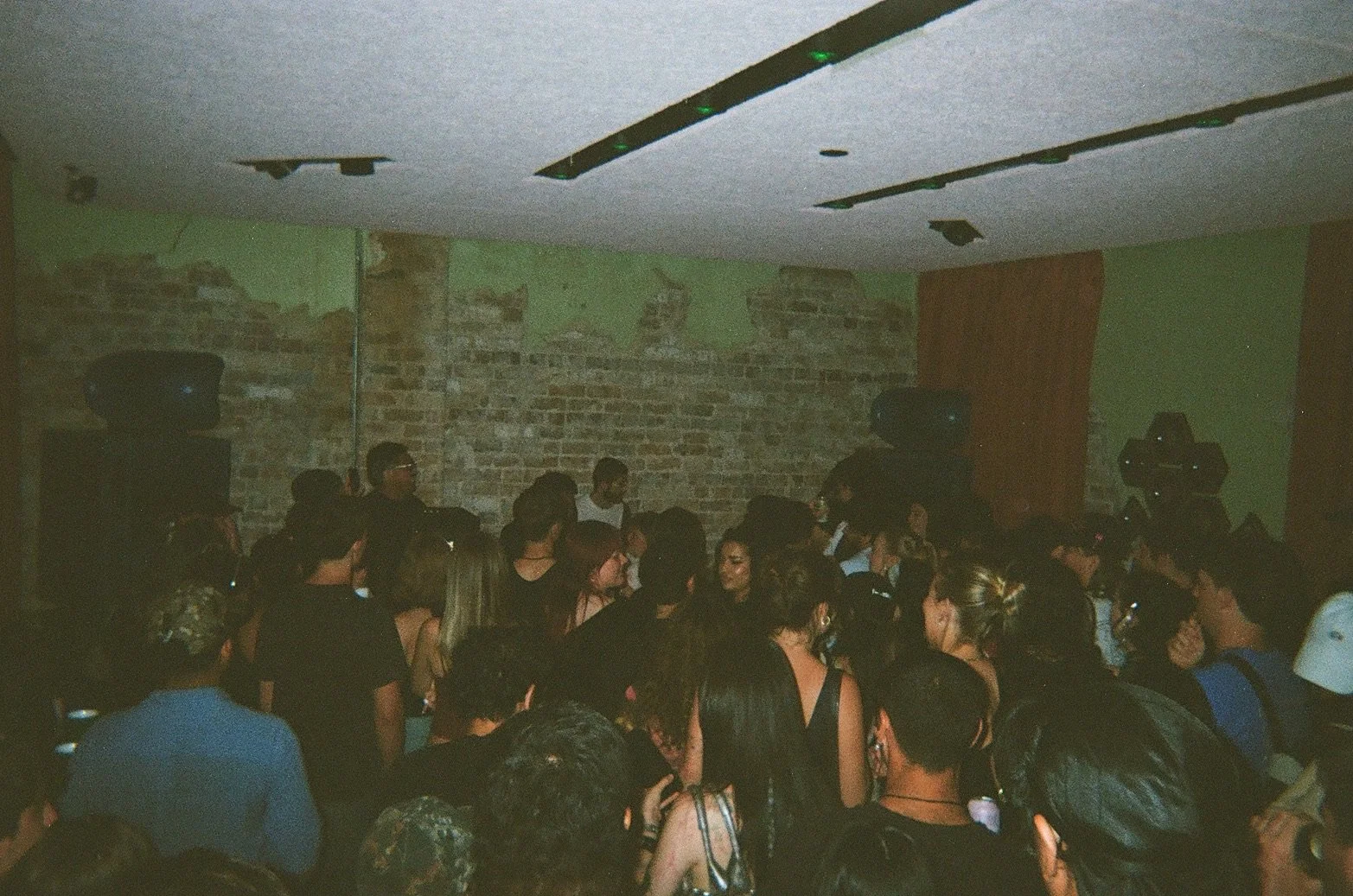 A crowded indoor concert scene with a brick wall backdrop and speakers on either side, filled with people socializing and enjoying music.