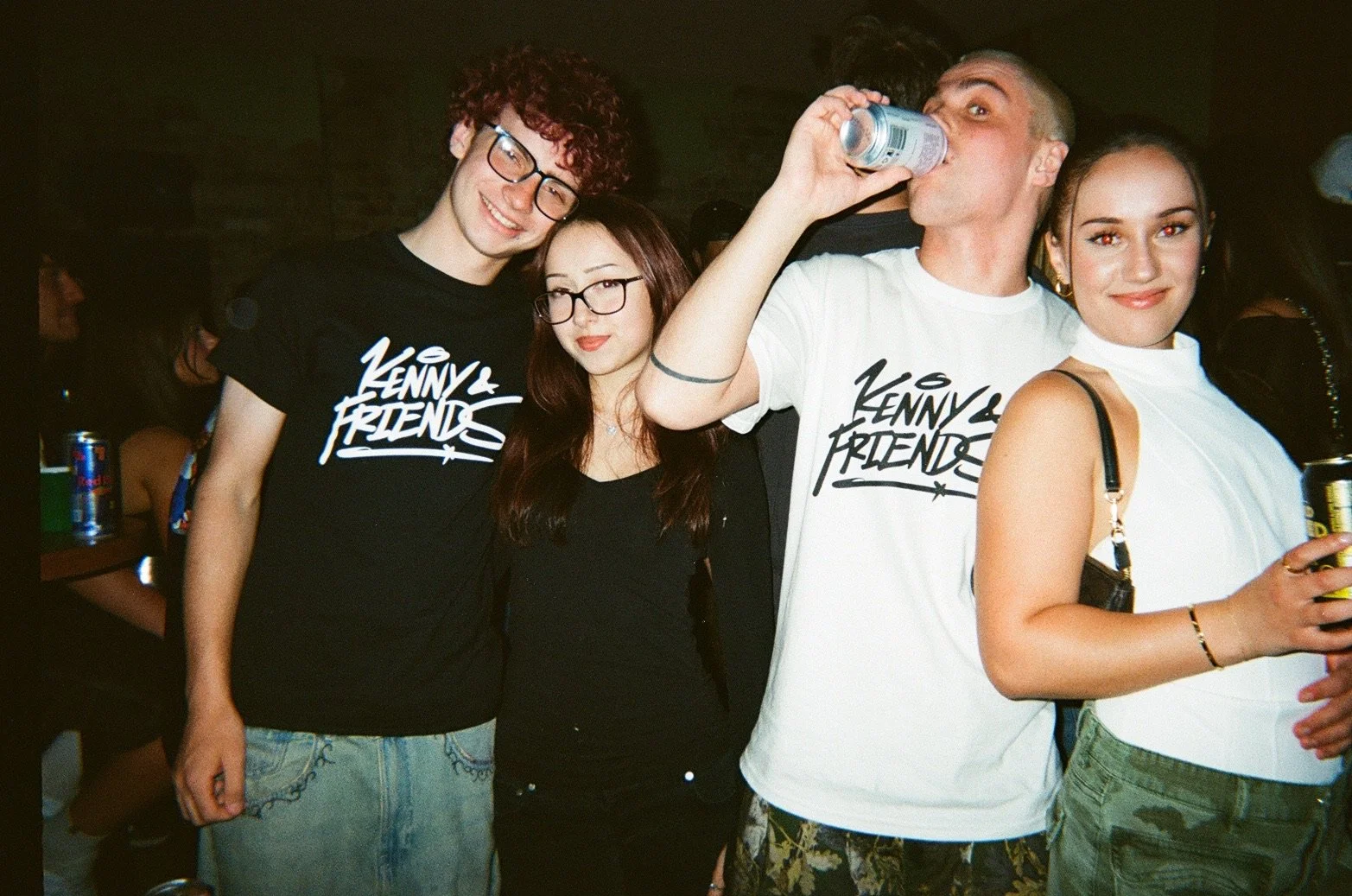 Group of four young people in casual attire at a party, two men and two women, some holding drinks, smiling and enjoying themselves.
