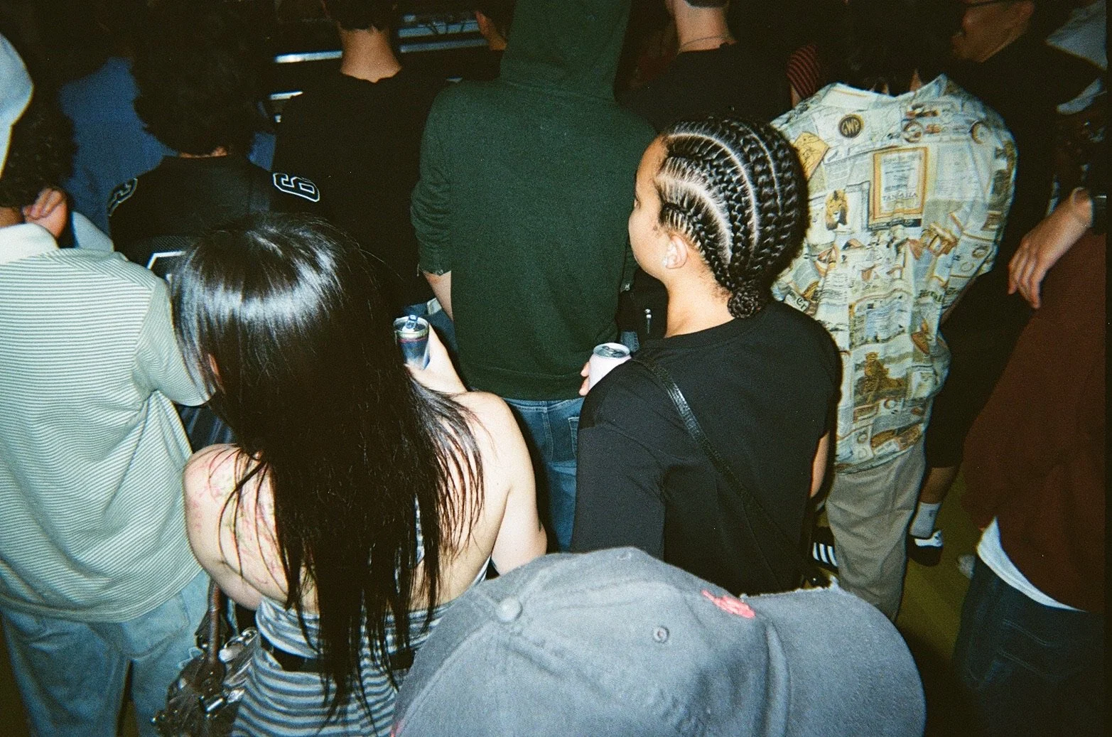Group of people standing closely together, some holding cans, with varied hairstyles and clothing, seen from behind in an indoor setting.