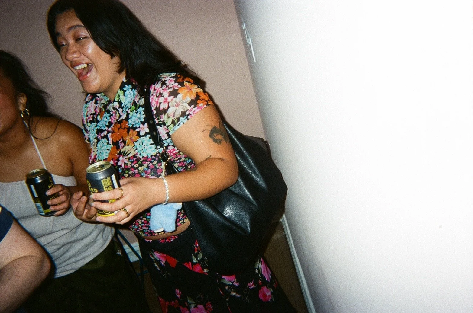 A woman with long dark hair wearing a colorful floral shirt and black skirt, holding a can of drink, smiling and laughing at a social gathering.