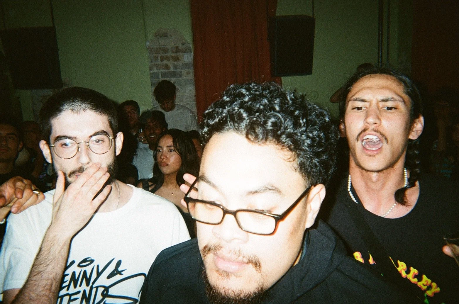 Group of young adults at a party or concert, some with glasses, hairstyles, and expressions indicating they are enjoying music or socializing.