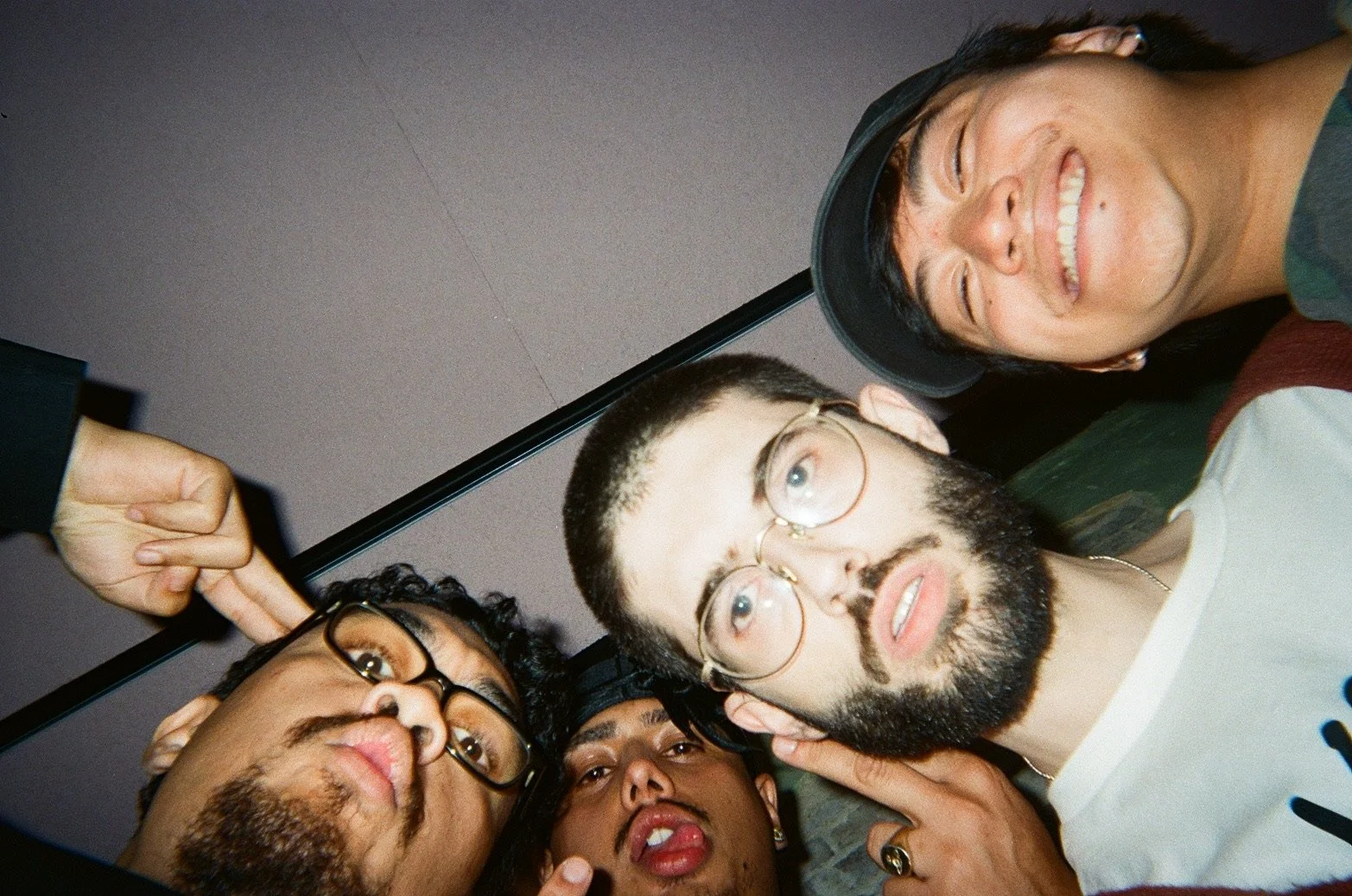 Group of four diverse friends in a selfie, smiling and making playful gestures, with a person wearing glasses and a gray shirt in the foreground.