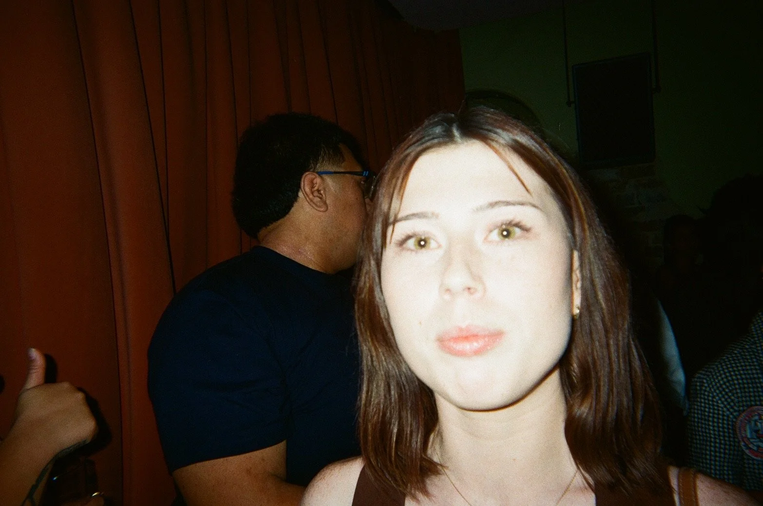 Close-up of a woman with light skin, brown hair, and green eyes looking at the camera. Behind her, a man with short dark hair, glasses, and a dark shirt is turned away from the camera, talking to someone out of frame. The background shows wooden wall