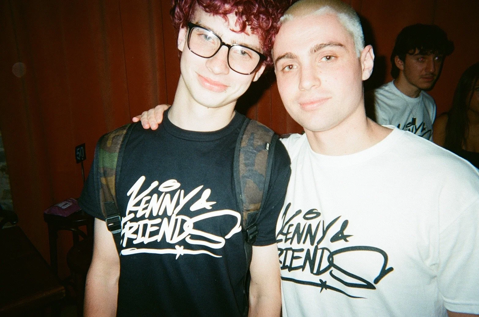 Two young men standing close together, smiling at the camera, wearing matching "Lenny & Friends" T-shirts. One has curly red hair, glasses, and a backpack; the other has bleached blond hair and no glasses.