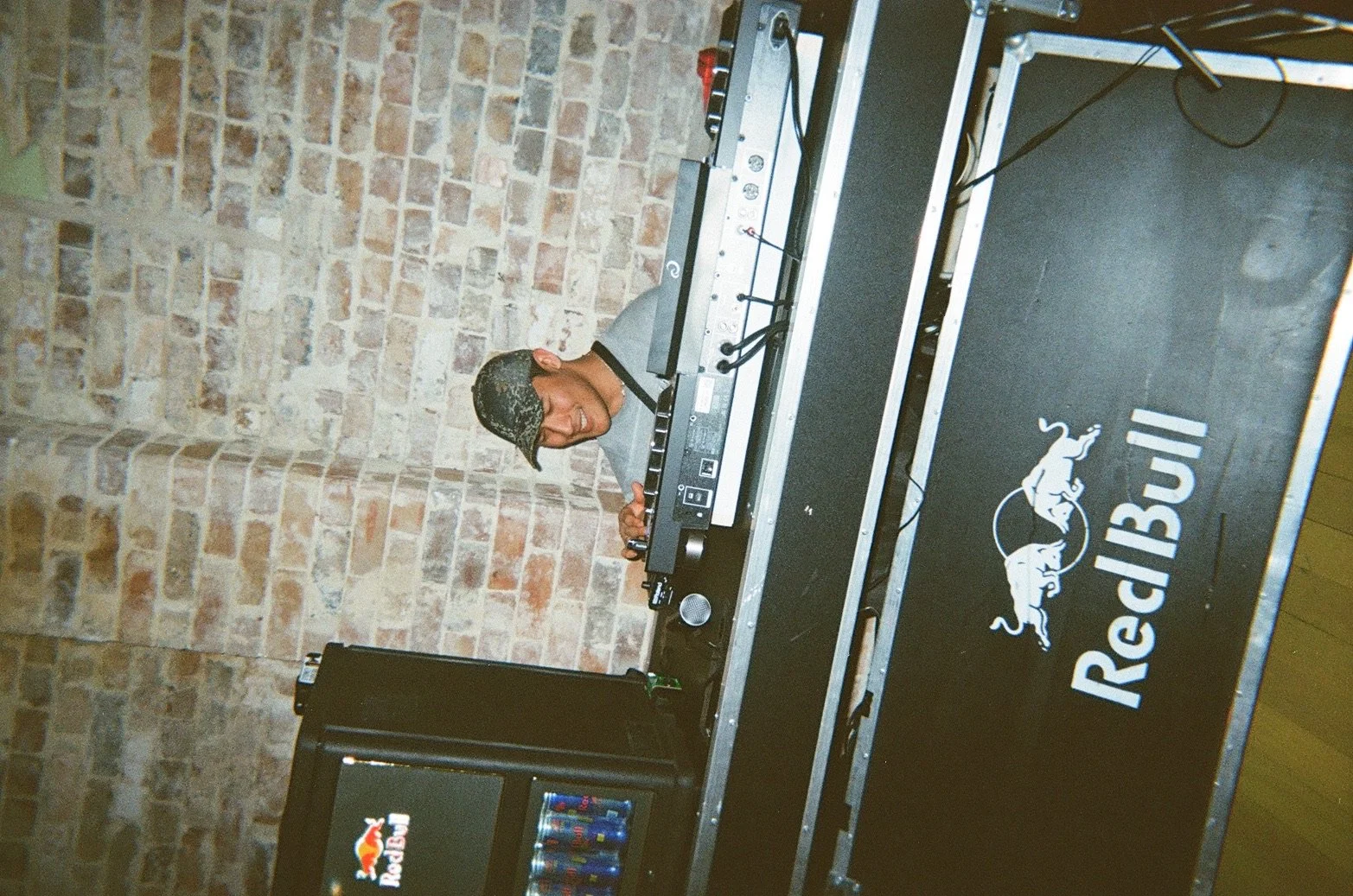 A young man wearing a camouflage cap and a gray shirt is DJing at a Red Bull event, standing behind a DJ booth with a laptop and audio equipment. The background features a brick wall.