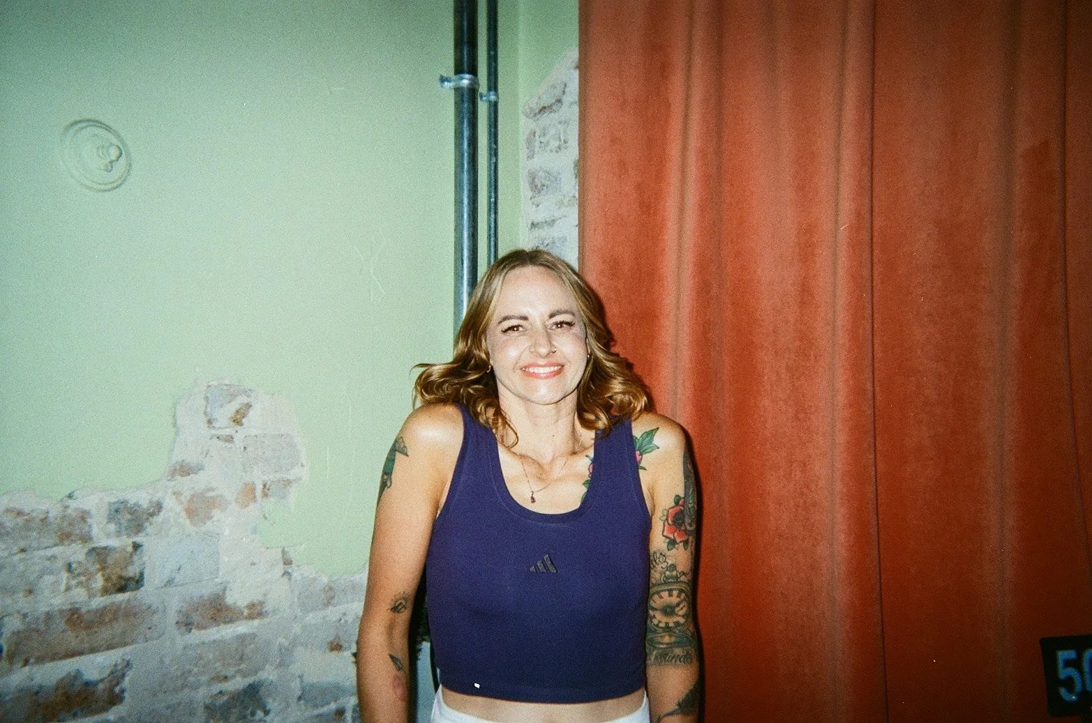A woman with shoulder-length red hair smiling, wearing a dark blue sleeveless top and white pants, standing in front of a wall with exposed brick and red curtains.