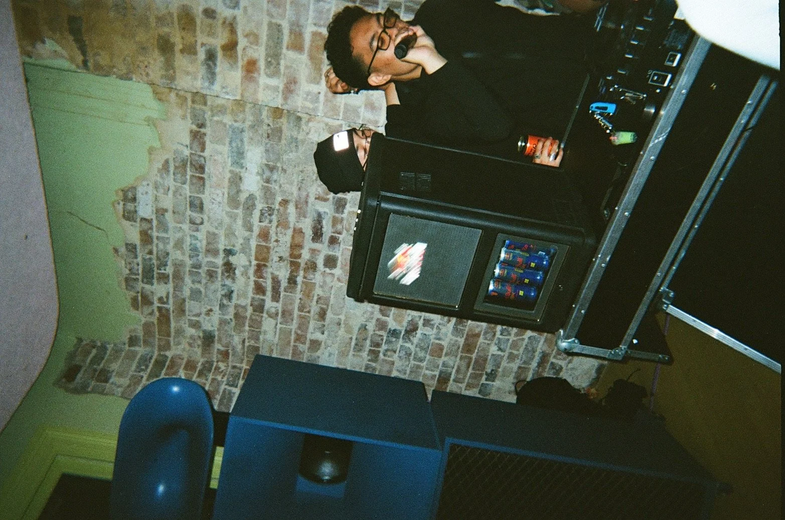 Two young men at a DJ booth in a room with brick walls, one wearing glasses, headphones, and black clothing, the other with a beanie, both standing behind a sound system with a small fridge with cans inside, and a speaker nearby.
