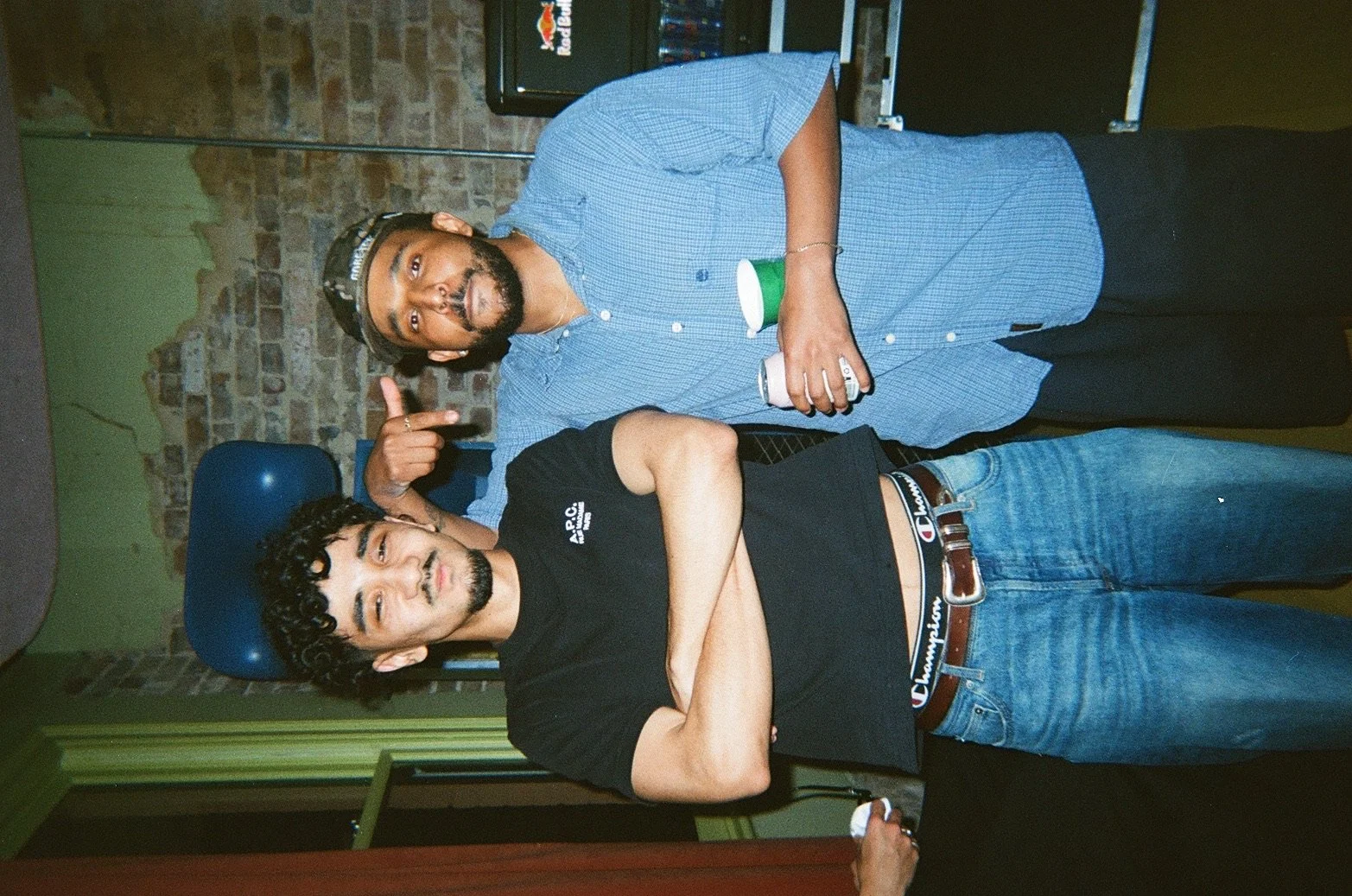 Two men standing together in a room with exposed brick wall background. The man on the left has curly dark hair, a beard, and is wearing a black T-shirt and blue jeans with a brown belt. The man on the right has a beard and mustache, is wearing a blu