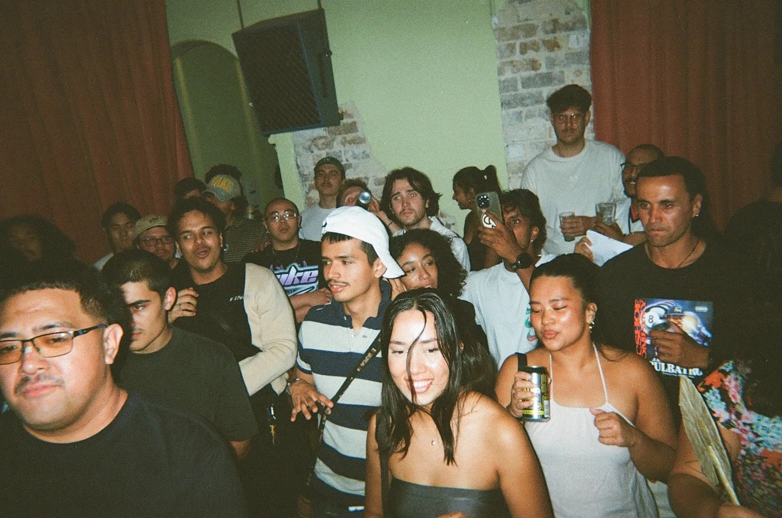A group of people at a party, with some holding drinks and taking photos, in a room with exposed brick and green walls.