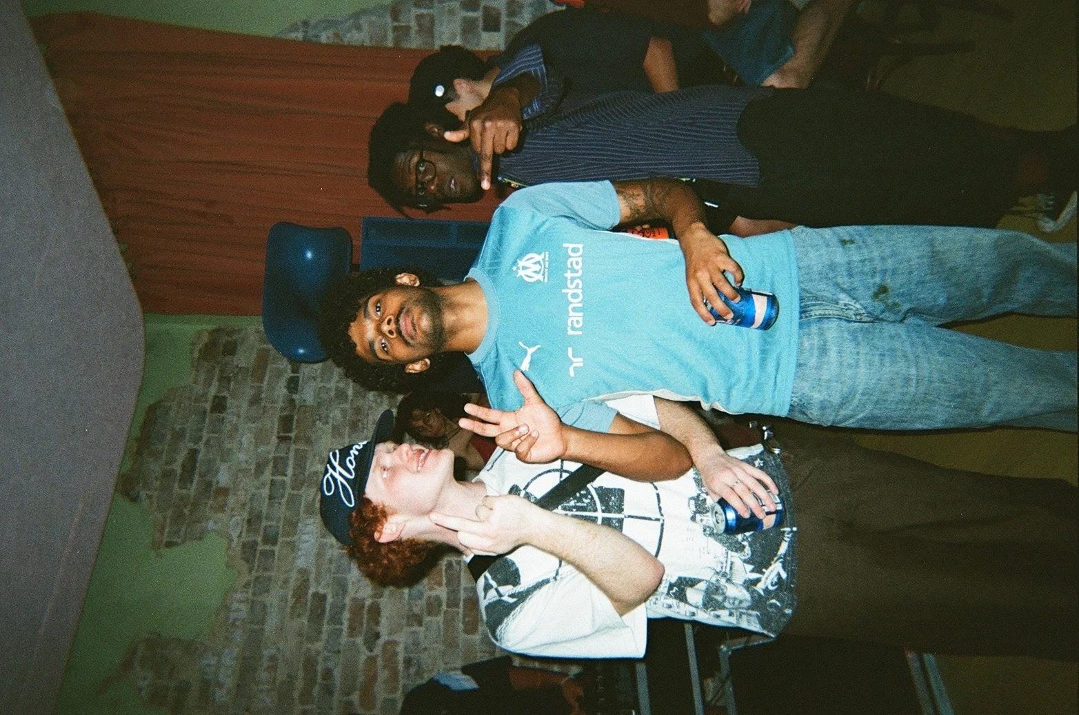Two young men at a social gathering, one with curly red hair wearing a black cap and a graphic T-shirt, making a peace sign with his hand. The other with glasses and black hair wearing a blue T-shirt and holding an open can, also making a peace sign.