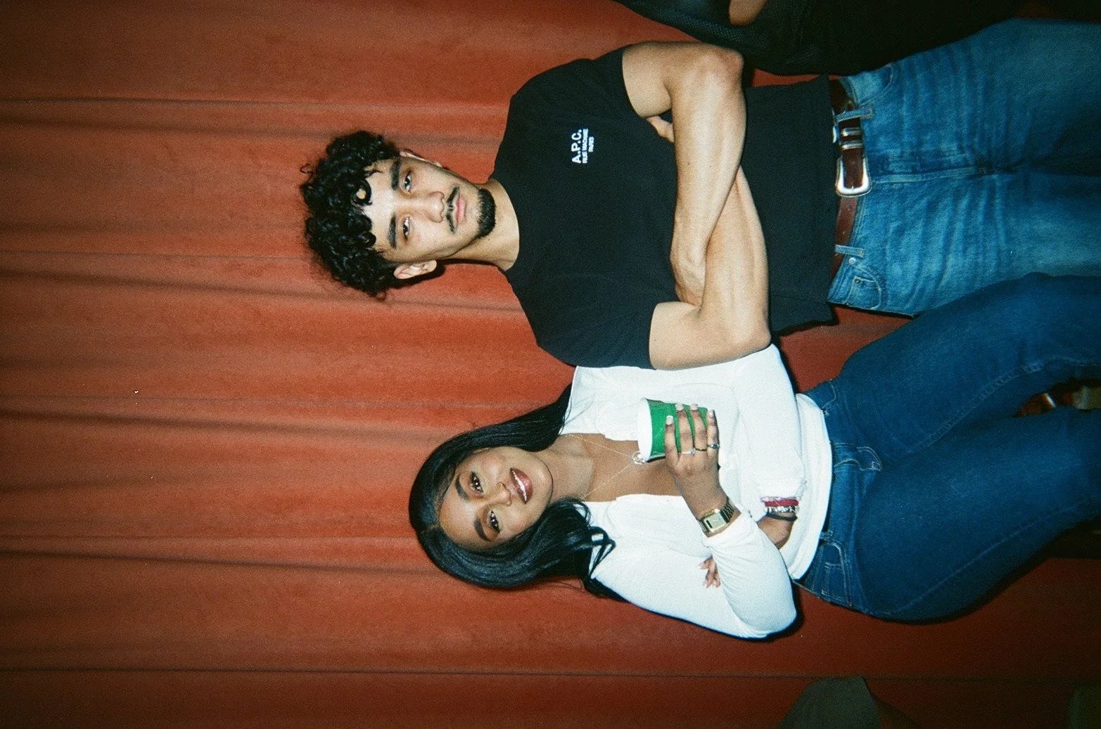 A man and a woman standing against a plain red curtain, posing for the photo. The man is wearing a black t-shirt and jeans, with arms crossed, looking at the camera. The woman is wearing a white top and jeans, holding a green cup, and also looking at