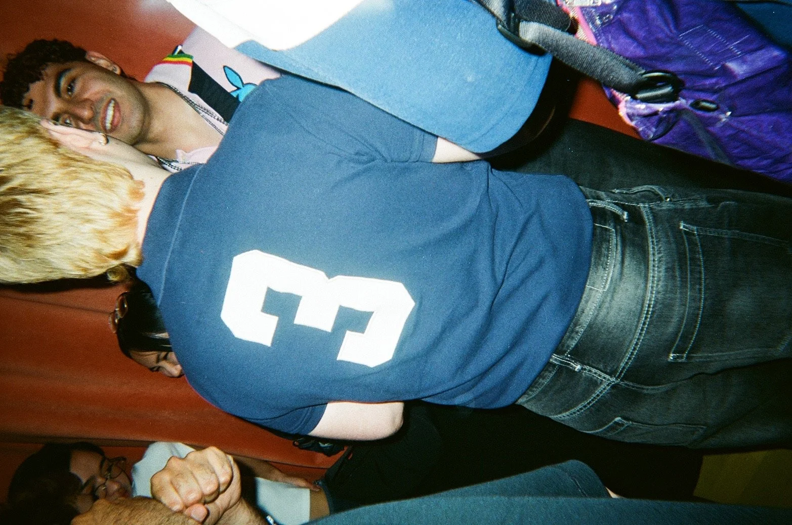 Two young men are close together, one with blond hair and the other with dark curly hair, engaging in conversation in what appears to be a social setting.