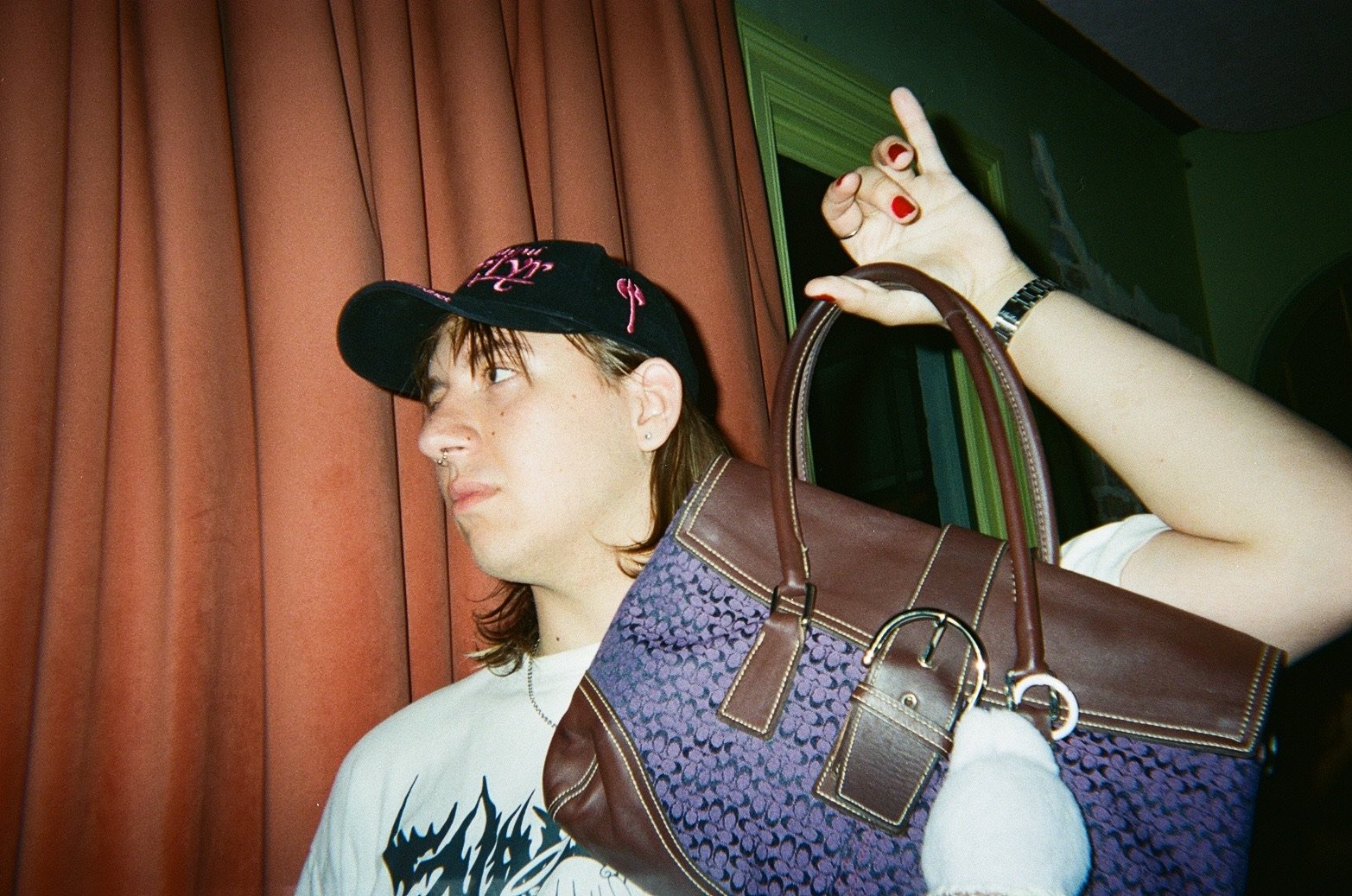 A young person with long hair, wearing a black baseball cap and a white T-shirt, holding a brown and purple handbag over their shoulder. They are standing in front of a reddish-brown curtain with a green door frame nearby.