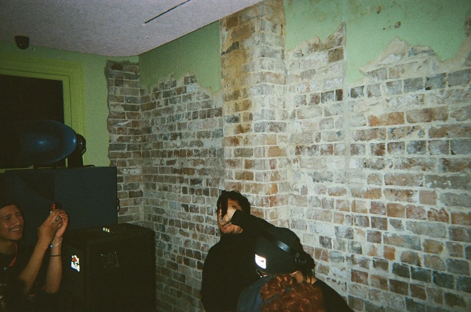 Group of people indoors with a brick wall in the background; one person is smiling, another is covering their face with a hand, a third person is wearing a black cap, and others are partially visible.