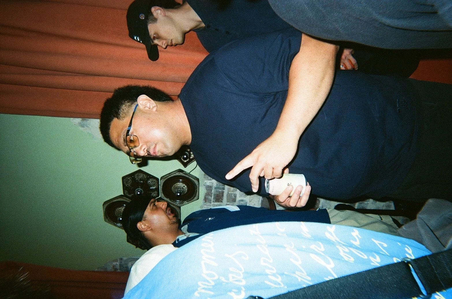 Three men, two standing and one lying down, are engaged in conversation in a room with green walls, a brick wall section, and speakers mounted on the wall.