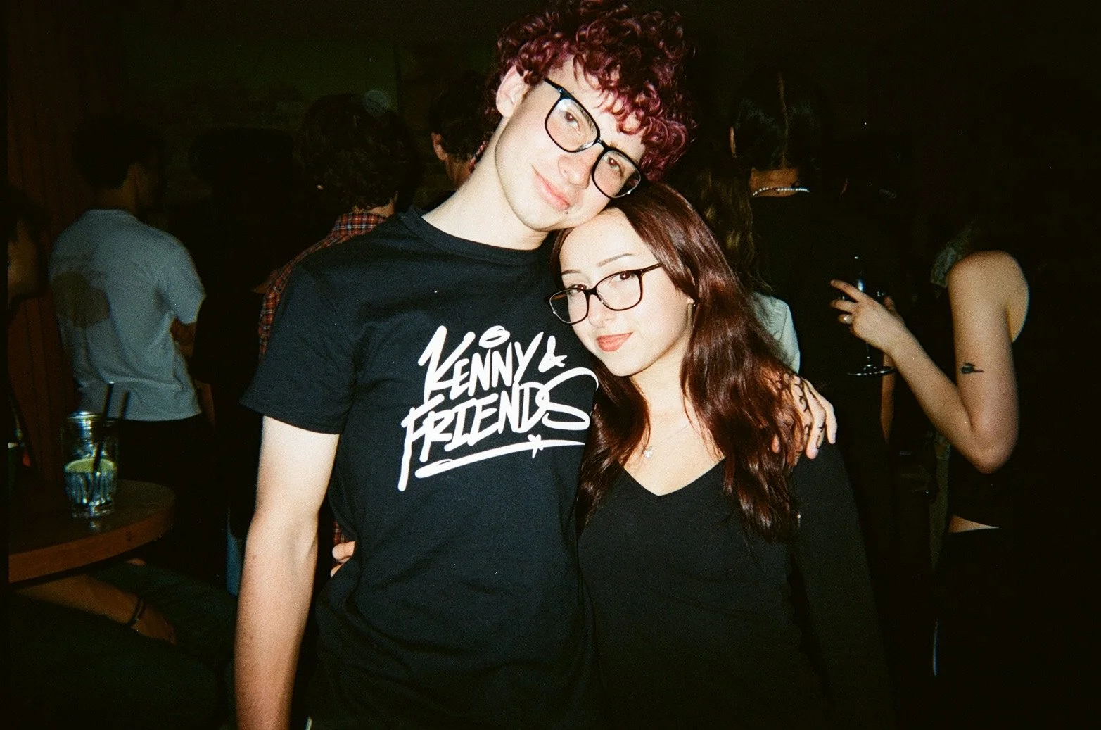 Young man with red curly hair and glasses wearing a black t-shirt that says 'Kenny & Friends', standing next to a young woman with long reddish-brown hair and glasses, both smiling at the camera, in a dimly lit social setting with other people in the