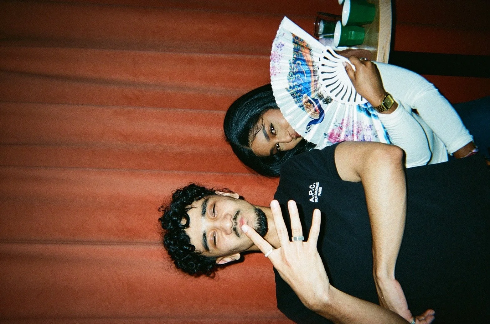 Two people sitting by a wooden wall, one holding a colorful fan partially covering their face, and the other making a peace sign with their hand. Both appear to be relaxed and casual.