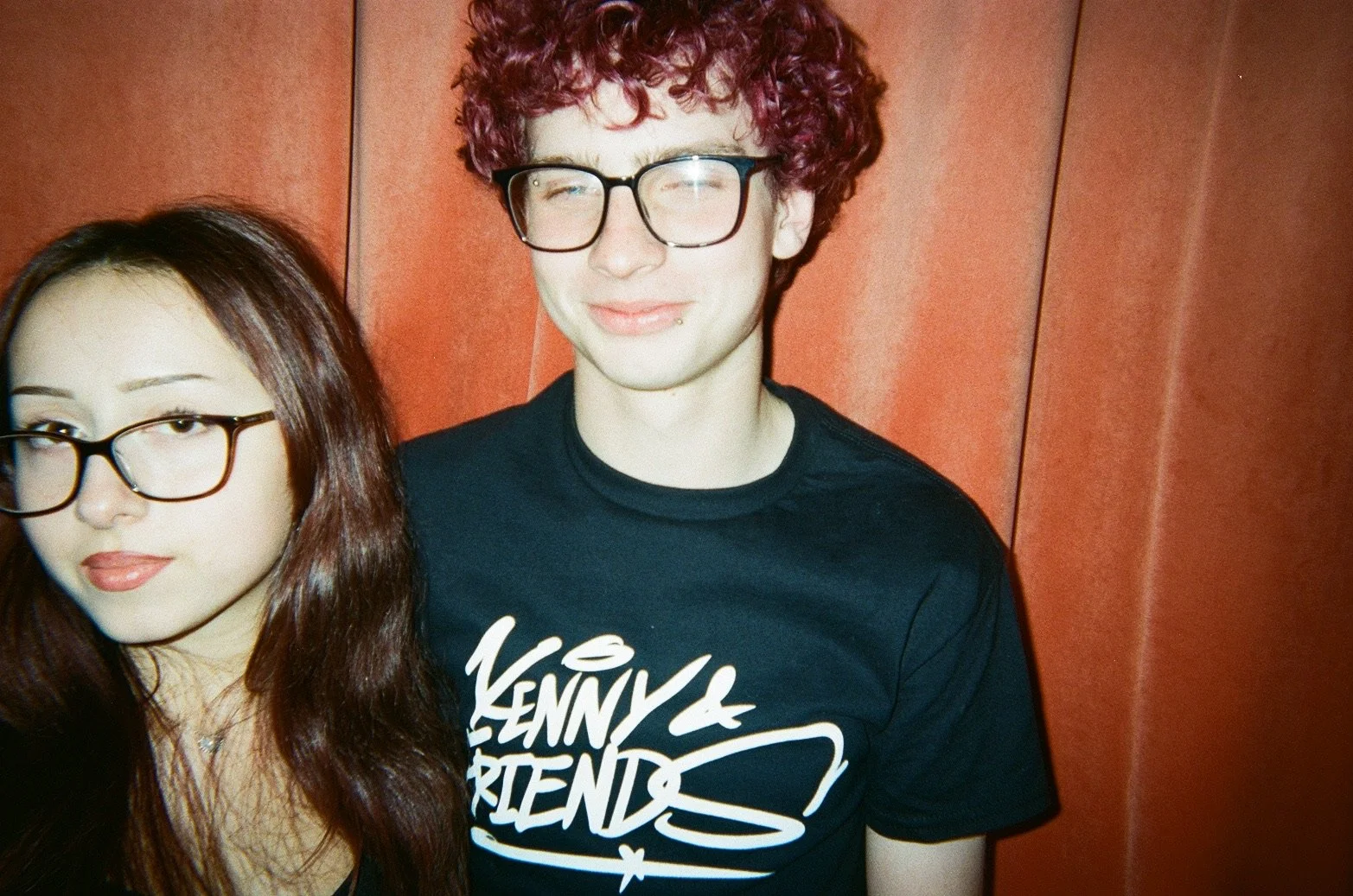 A young man with curly red hair, glasses, and a lip piercing is smiling with eyes closed, wearing a black T-shirt with white text that reads 'Venny & Friends'. Part of a young woman with glasses, long dark hair, and makeup is visible on the left side