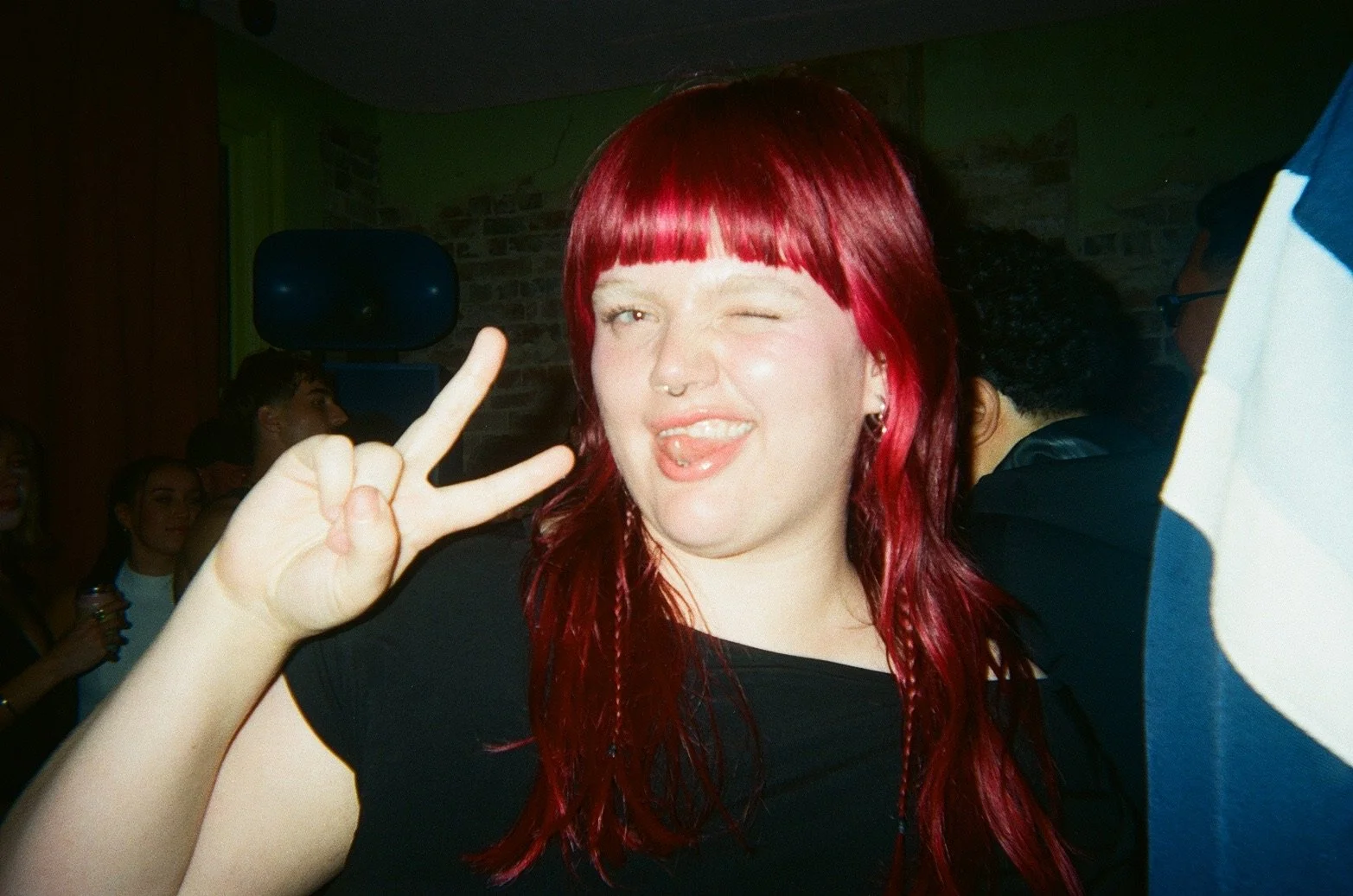 A young woman with red hair making a peace sign and sticking out her tongue in a social setting. She has a tongue piercing and a septum ring, wearing a black top.