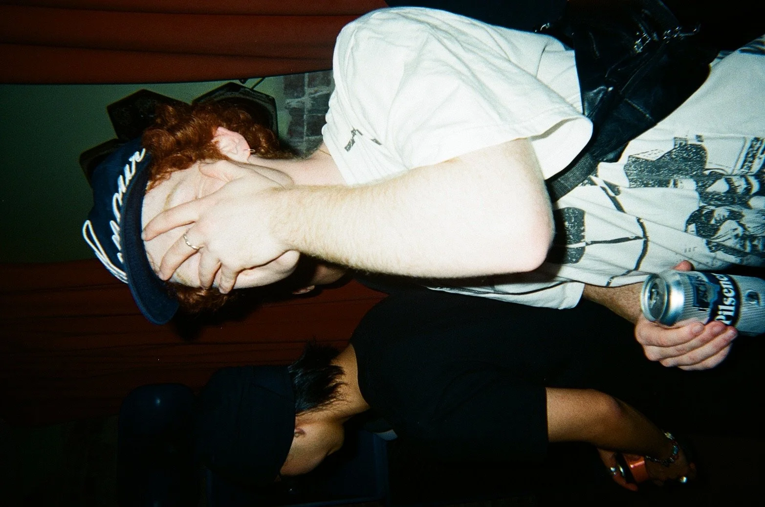 A young man with red hair and a cap is covering his face with his hand, holding a can of beer in his other hand, standing next to a woman with dark hair and a black shirt, against a background with a red curtain.