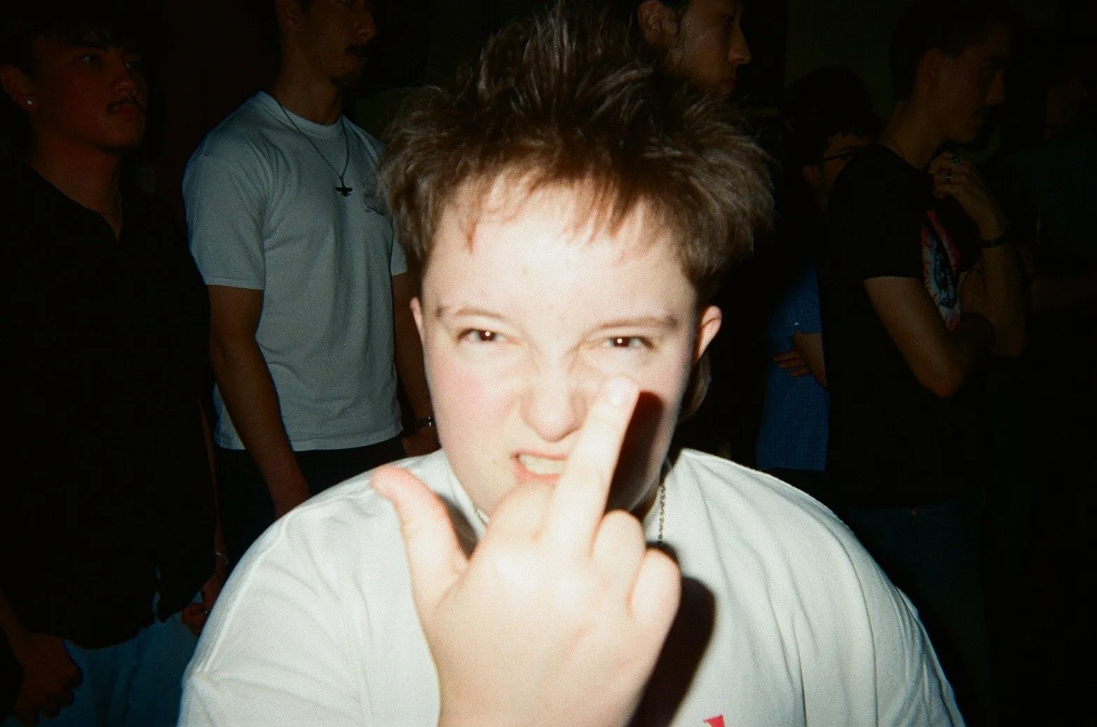 A person with short dark hair making an angry or defiant face, holding up their middle finger directly in front of the camera. Several other people are visible in the background, standing close together in a dimly lit environment.