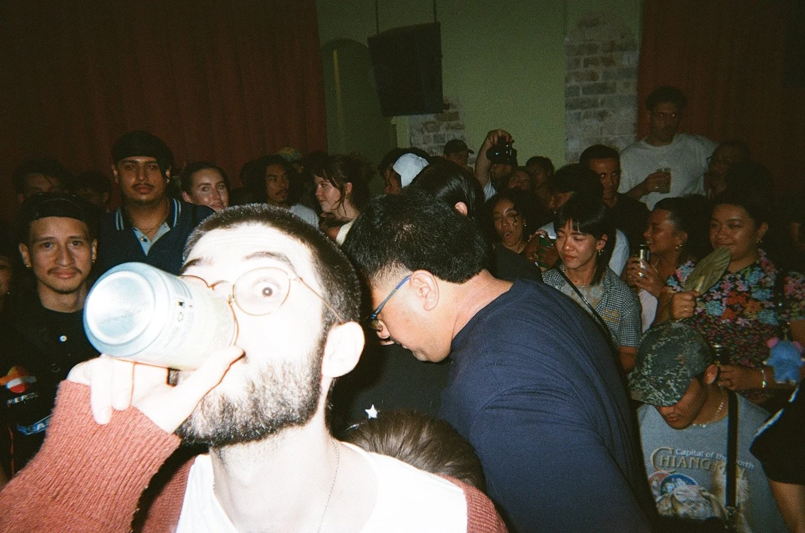 A crowded indoor party scene with diverse people, some taking photos and others talking, one person drinking from a can in the foreground.