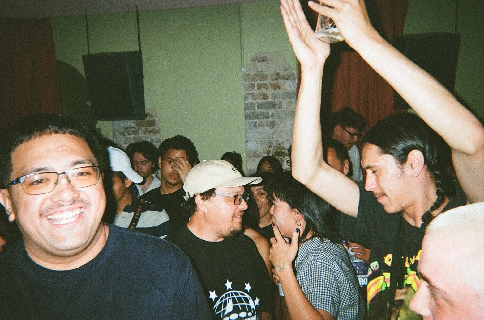 People dancing and socializing at a party, with one person raising a glass, in a room with exposed brick walls and green paint.