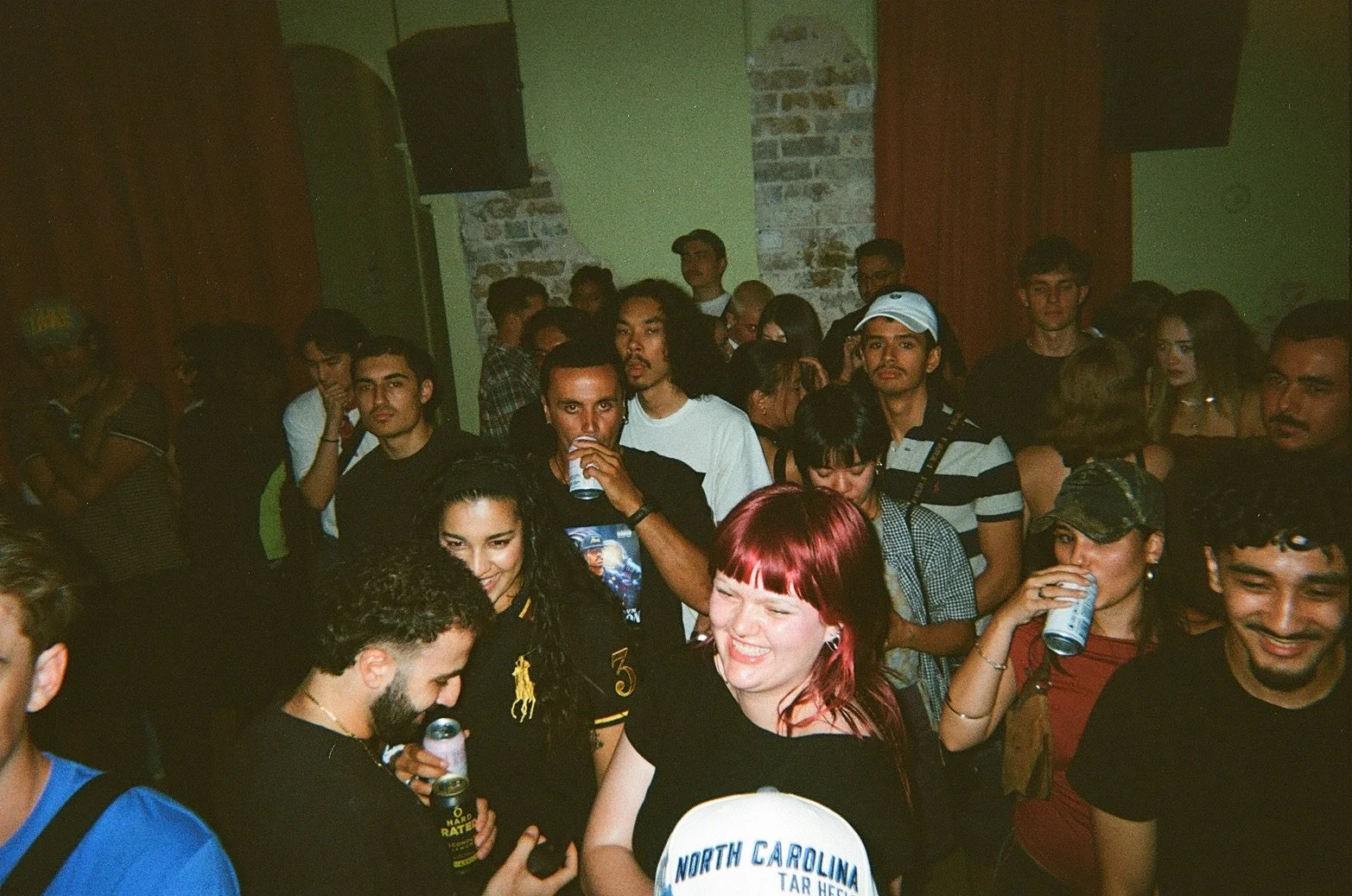 Group of young people at a party or concert, smiling and drinking, in a dimly lit indoor space with exposed brick walls.