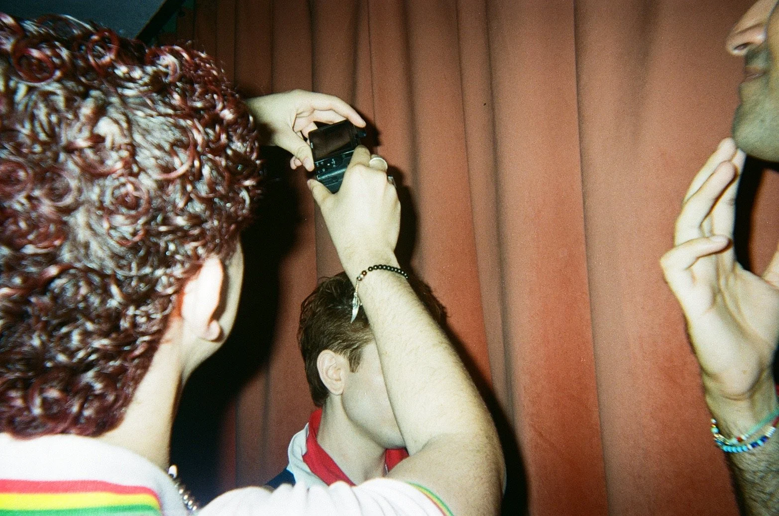 Two people taking photos of a person in front of a red curtain. One person has curly red hair and is holding a camera, while the other has short hair and is making an peace sign with their fingers.