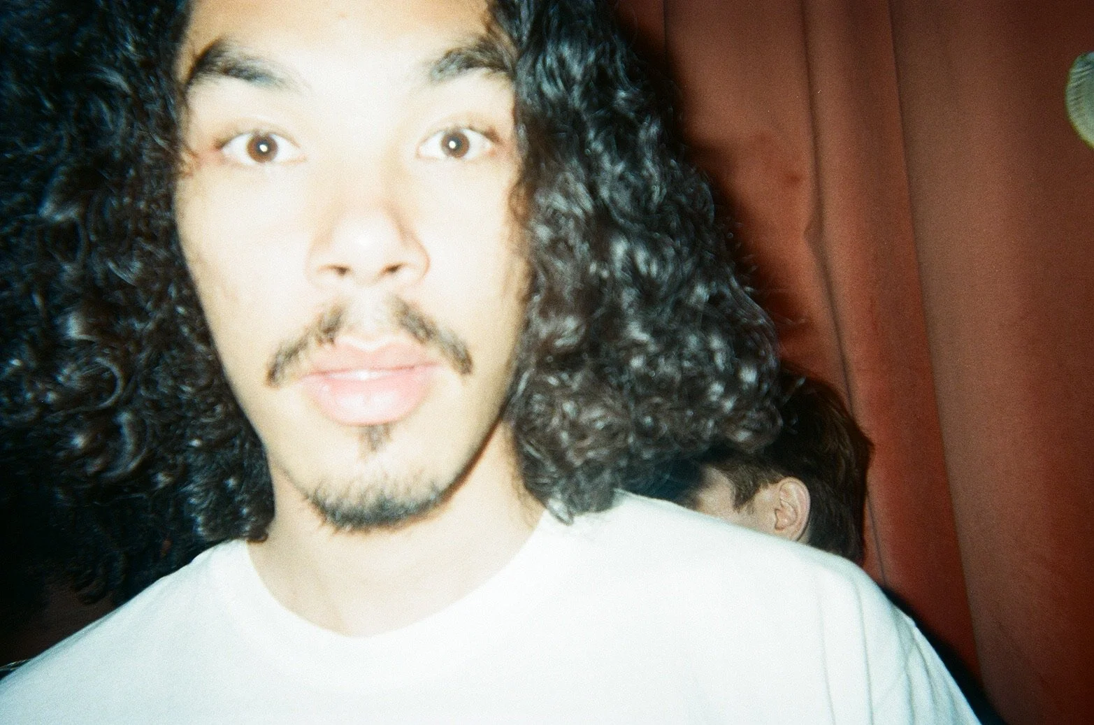 A close-up of a person with long curly black hair, a mustache, and a goatee, wearing a white shirt, looking directly at the camera.