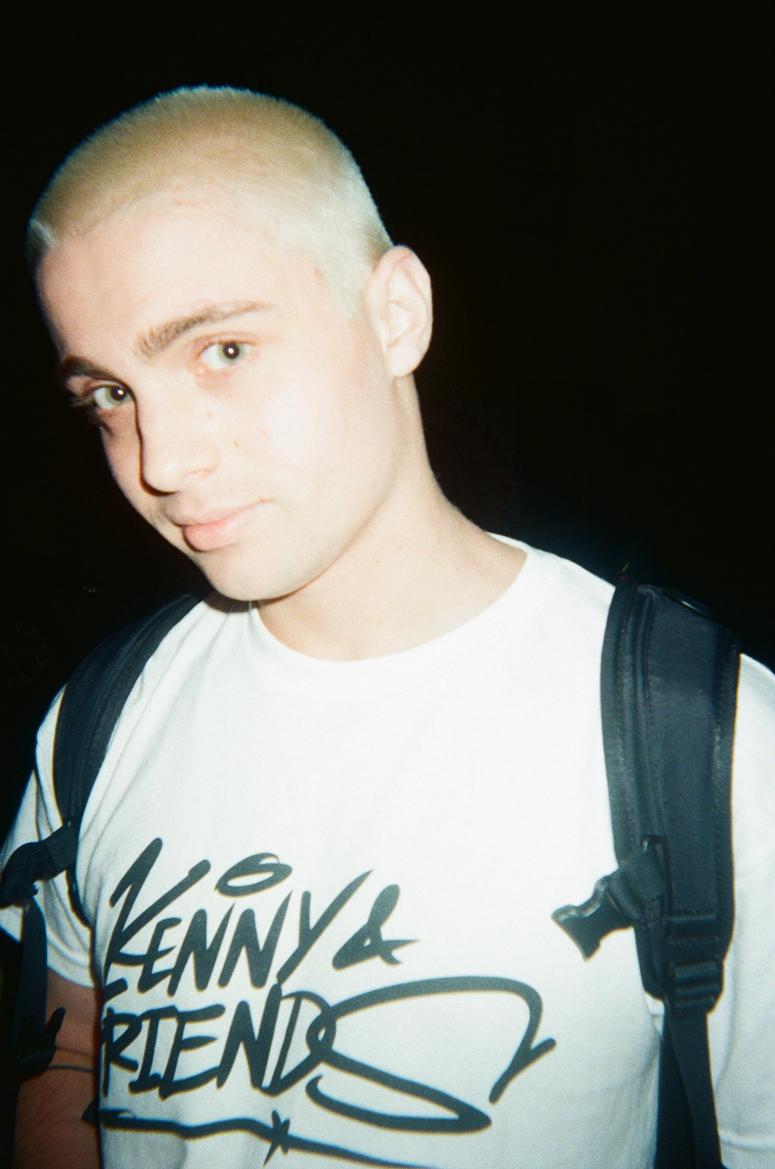 A young man with closely cropped blond hair, wearing a white t-shirt with black text and carrying a black backpack, looking at the camera with a slight smile.