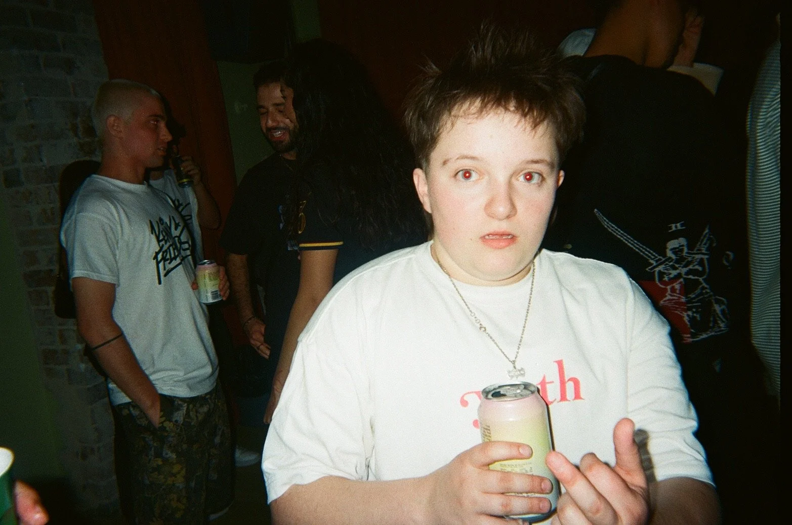 A young person with short dark hair holding a can, standing in a crowd at a party or social gathering, with multiple people in the background.