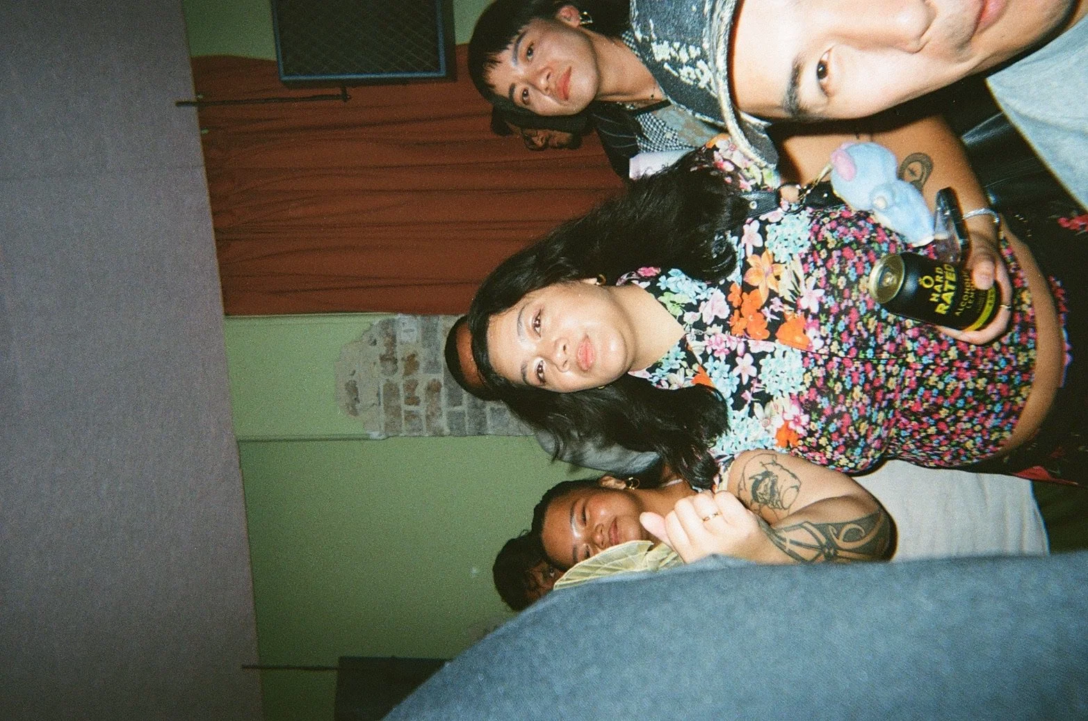 A group of friends sitting together indoors, some holding drinks, with a brick wall and curtain in the background