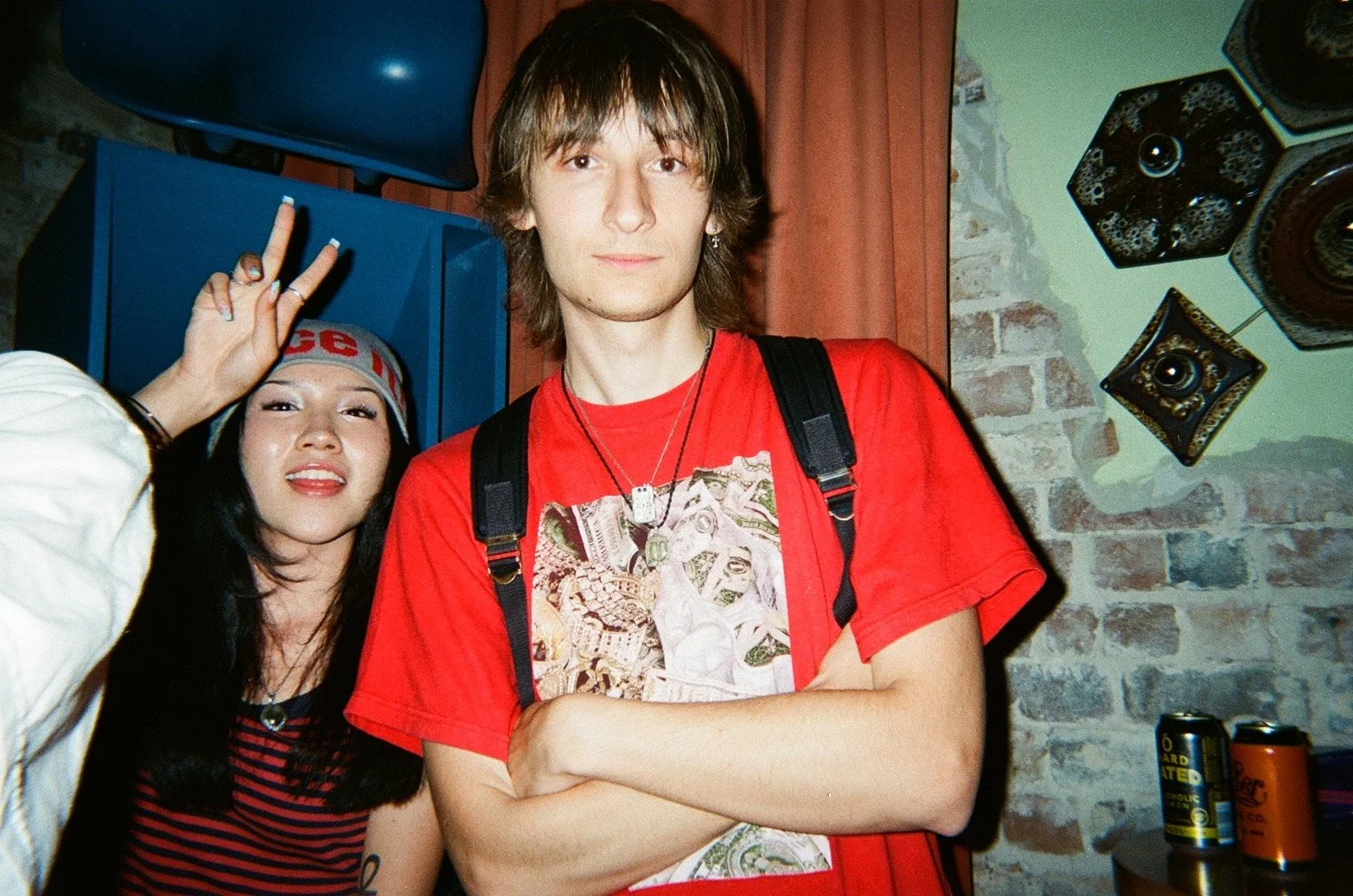 Two young people smiling and posing for a photo. The person in the foreground has long brown hair, wearing a red t-shirt with a graphic design, and crossed arms. The person in the background is wearing a striped red and black shirt, a cap, and is hol