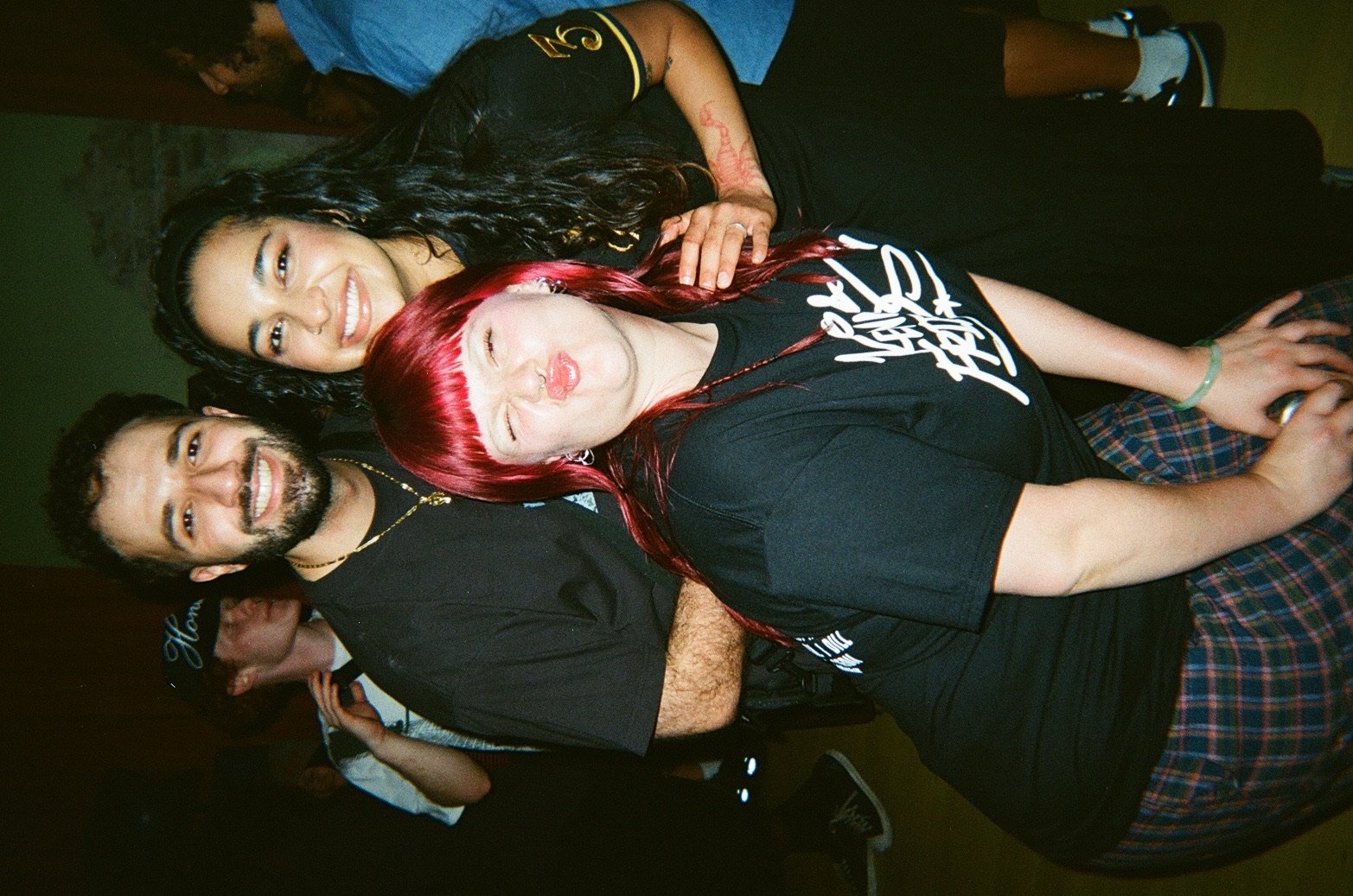 Three friends sitting closely together, smiling, with two women in front and a man behind them. The woman in the front has bright red hair, the woman in the middle has long curly black hair, and the man has a beard and short dark hair.