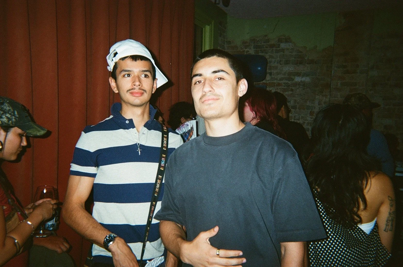 Two young men are posing for a photo at a social gathering. The man in the foreground is wearing a dark gray t-shirt and gestures with his right hand over his stomach. The man behind him is wearing a navy and white striped polo shirt, a white cap wor