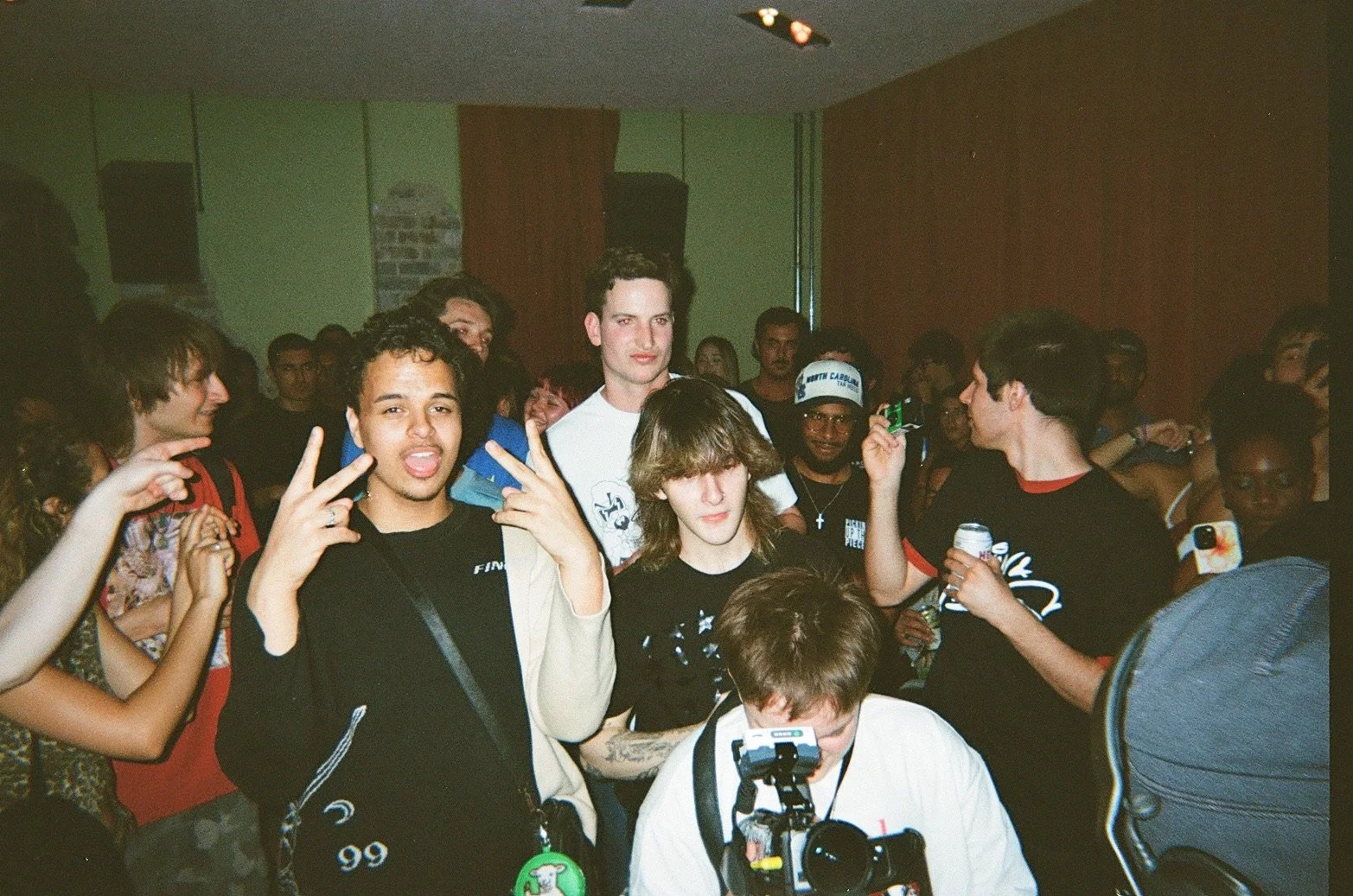 Group of young people at a crowded event, with some posing and holding drinks, and one person taking a photo with a camera.