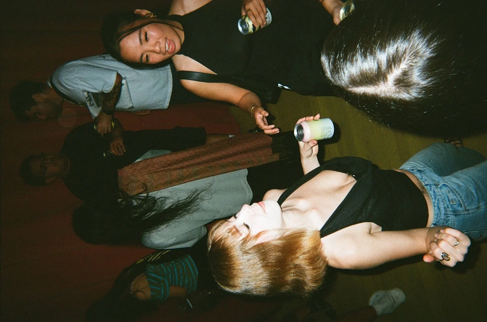Group of people at a party, some dancing and holding beverages, with a focus on two women in the foreground lying on the floor, one with blonde hair and the other with dark hair.