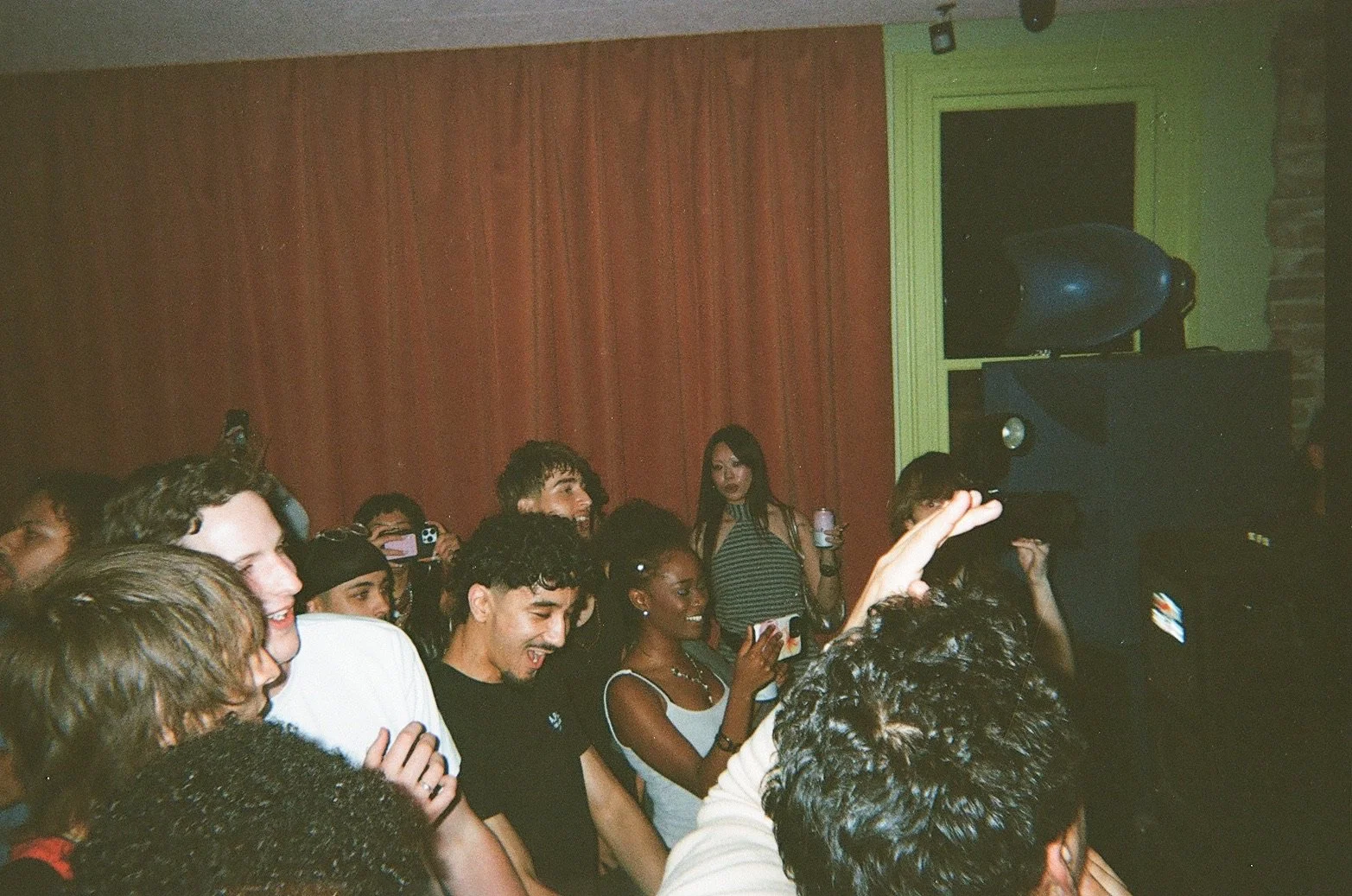 People dancing and enjoying music at a party with a red wall, green window frame, and a speaker in the background.