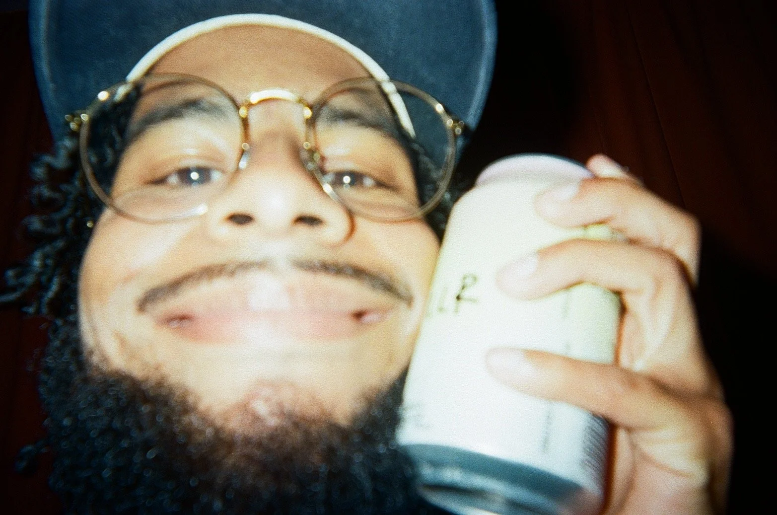 Close-up of a smiling man with glasses and a beard, wearing a cap, holding a can of beer near his face.