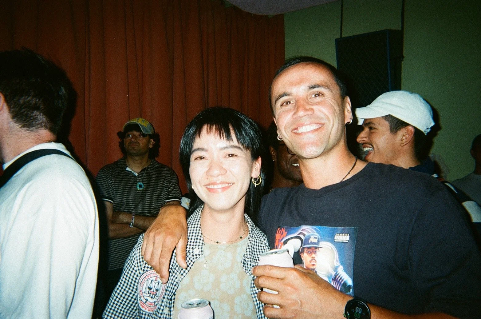 Two people smiling with arms around each other at a social event, with other attendees in the background.