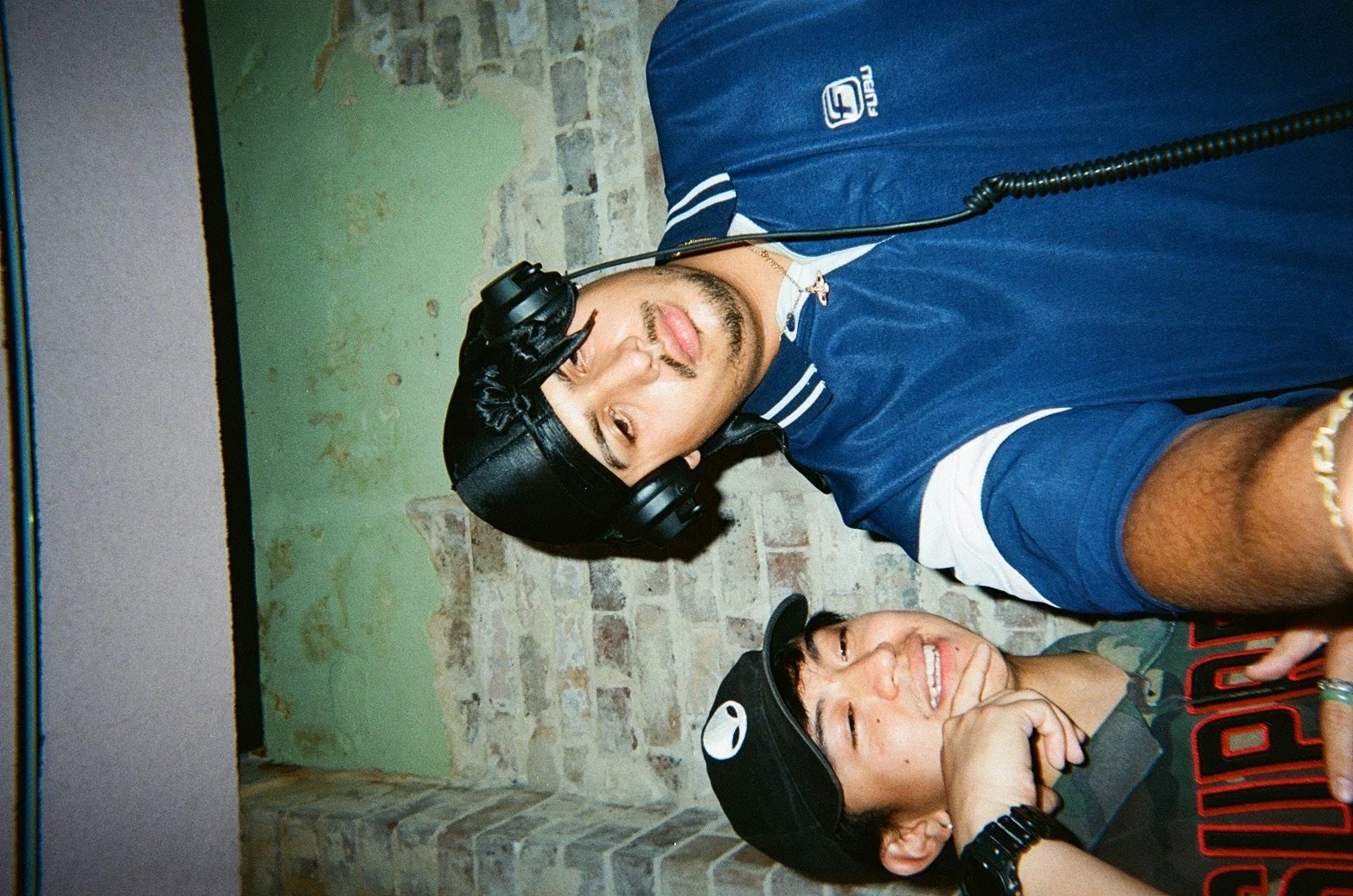 Two young men taking a selfie together, one wearing a black cap and headphones, the other wearing a black cap and a watch, both smiling and leaning against a brick wall.