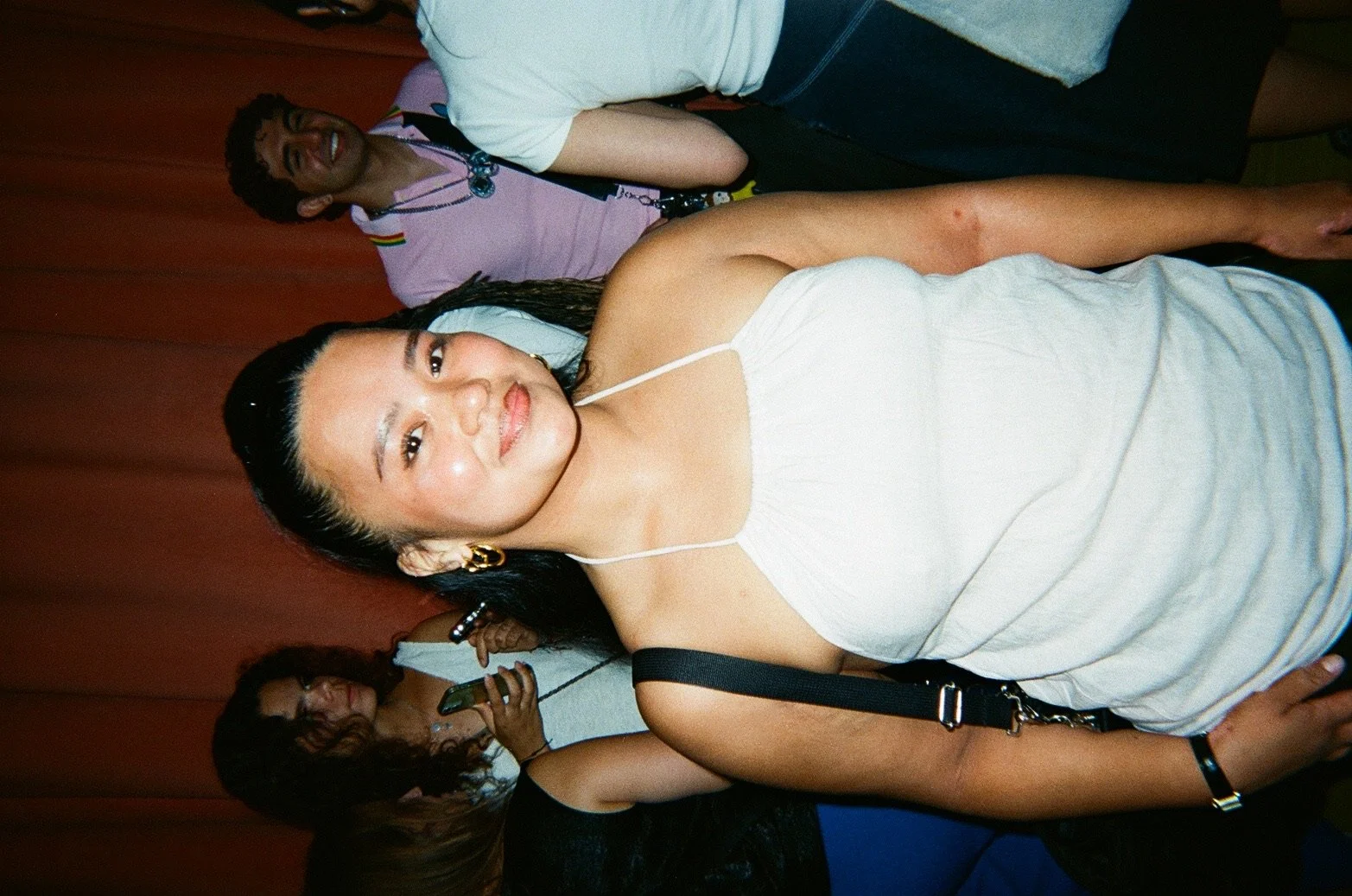A young woman with black hair in a white strap top and gold earrings, smiling at the camera, standing in front of a red curtain with other people in the background.