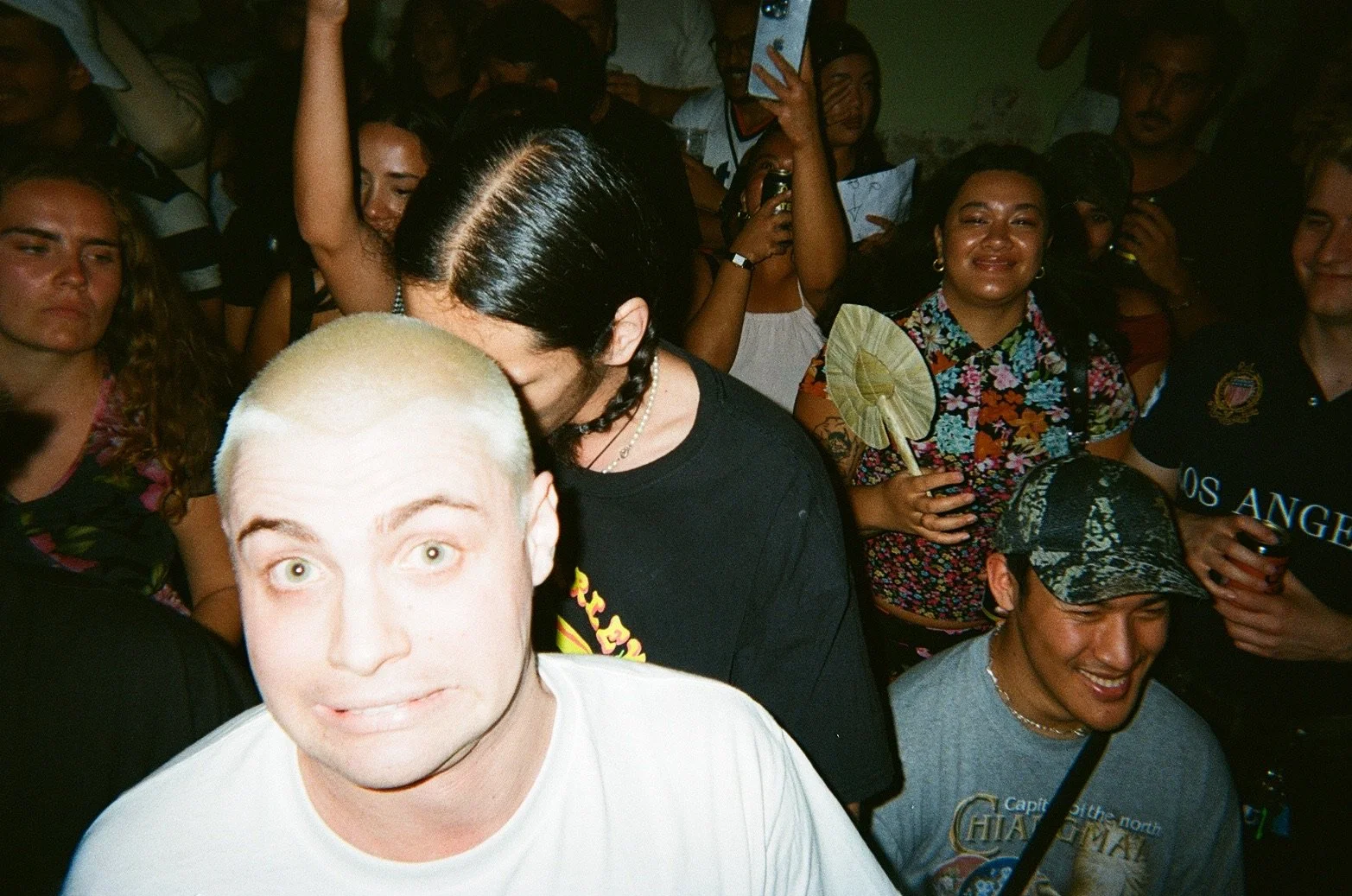 A crowded indoor party scene with diverse people, some smiling and holding drinks, and one person with bleached hair in the foreground.