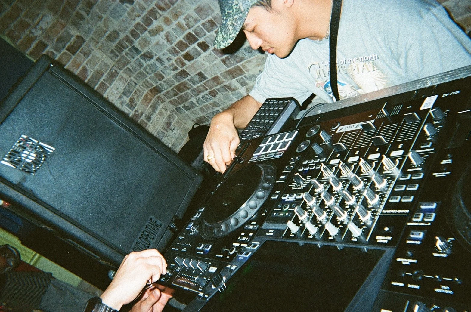 A DJ is mixing music on a professional DJ controller with a laptop, in front of a brick wall.